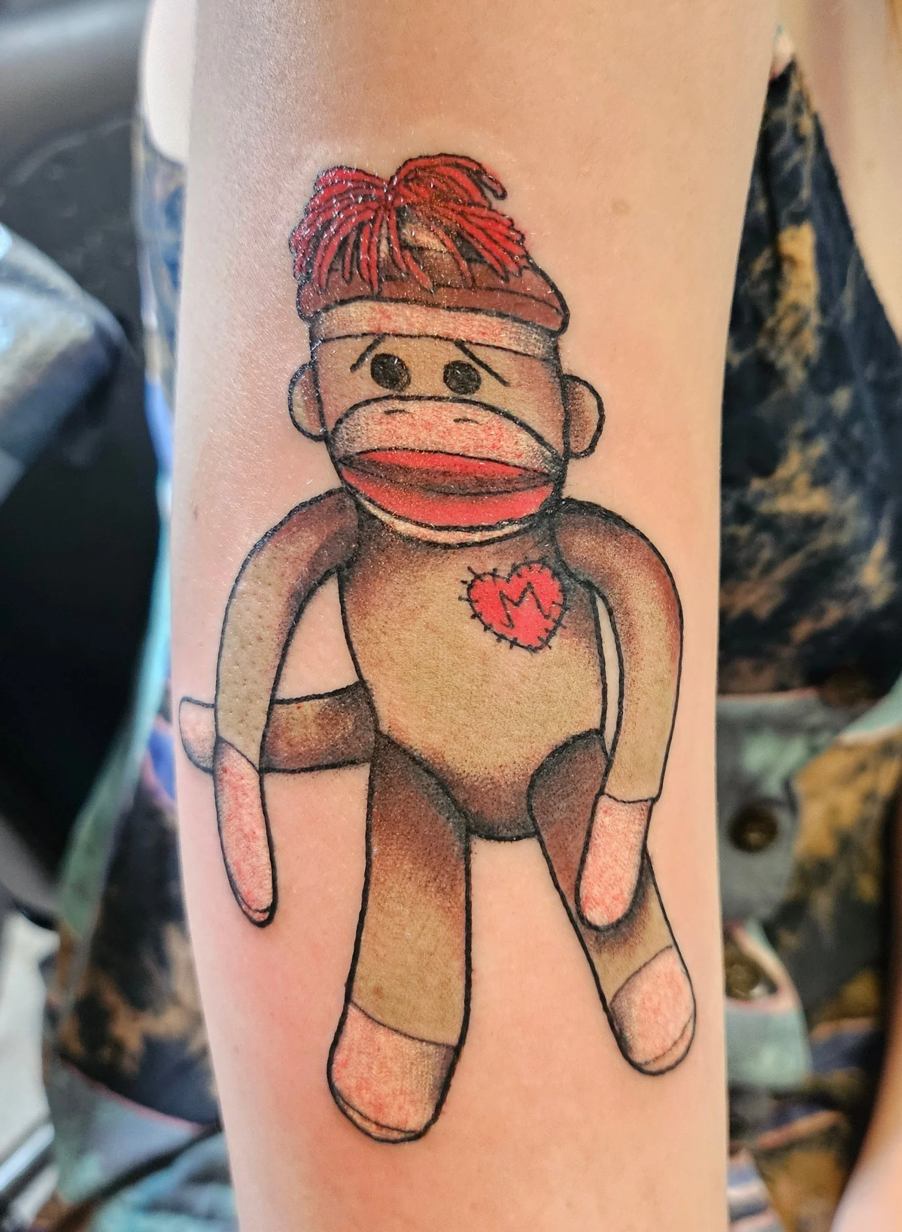 Tattoo of a monkey with a red mohawk, wearing a headband, gloves, and socks, with a heart and letter 'M' on its chest.
