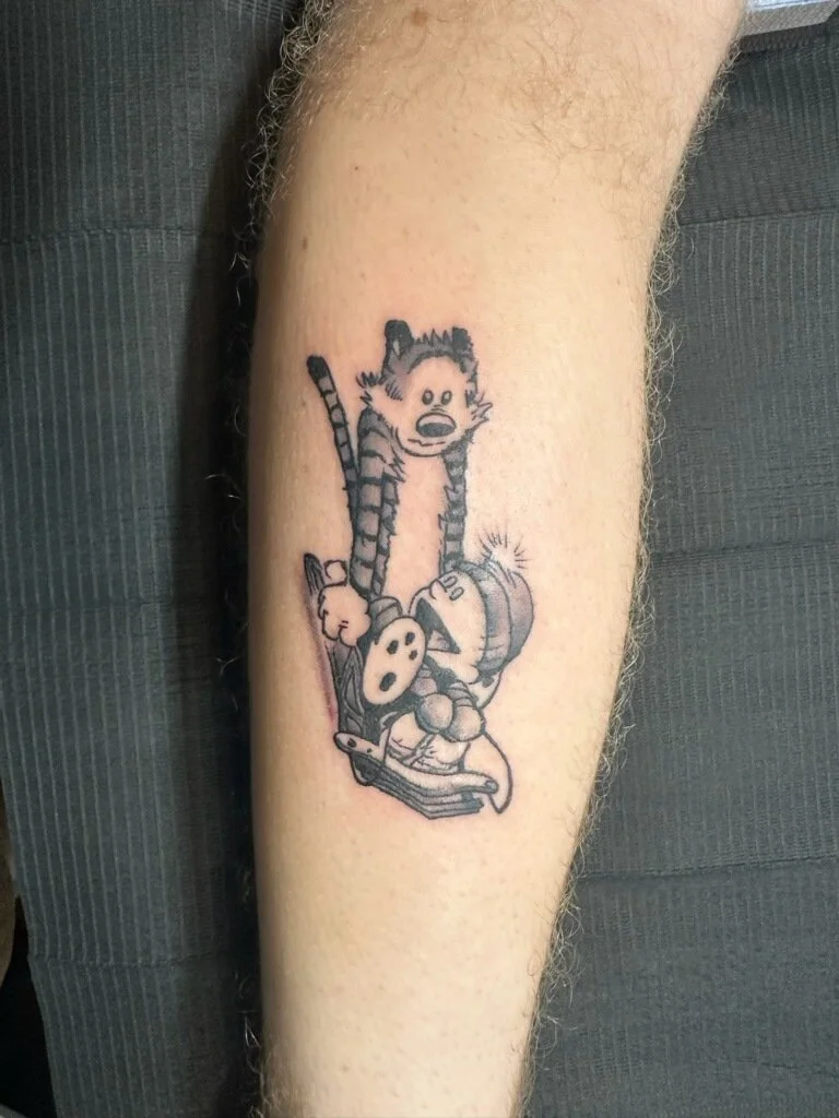 Tattoo of the comic strip character Calvin from Calvin and Hobbes, sitting on a couch, holding a game controller, with Hobbes standing behind him with a surprised expression.