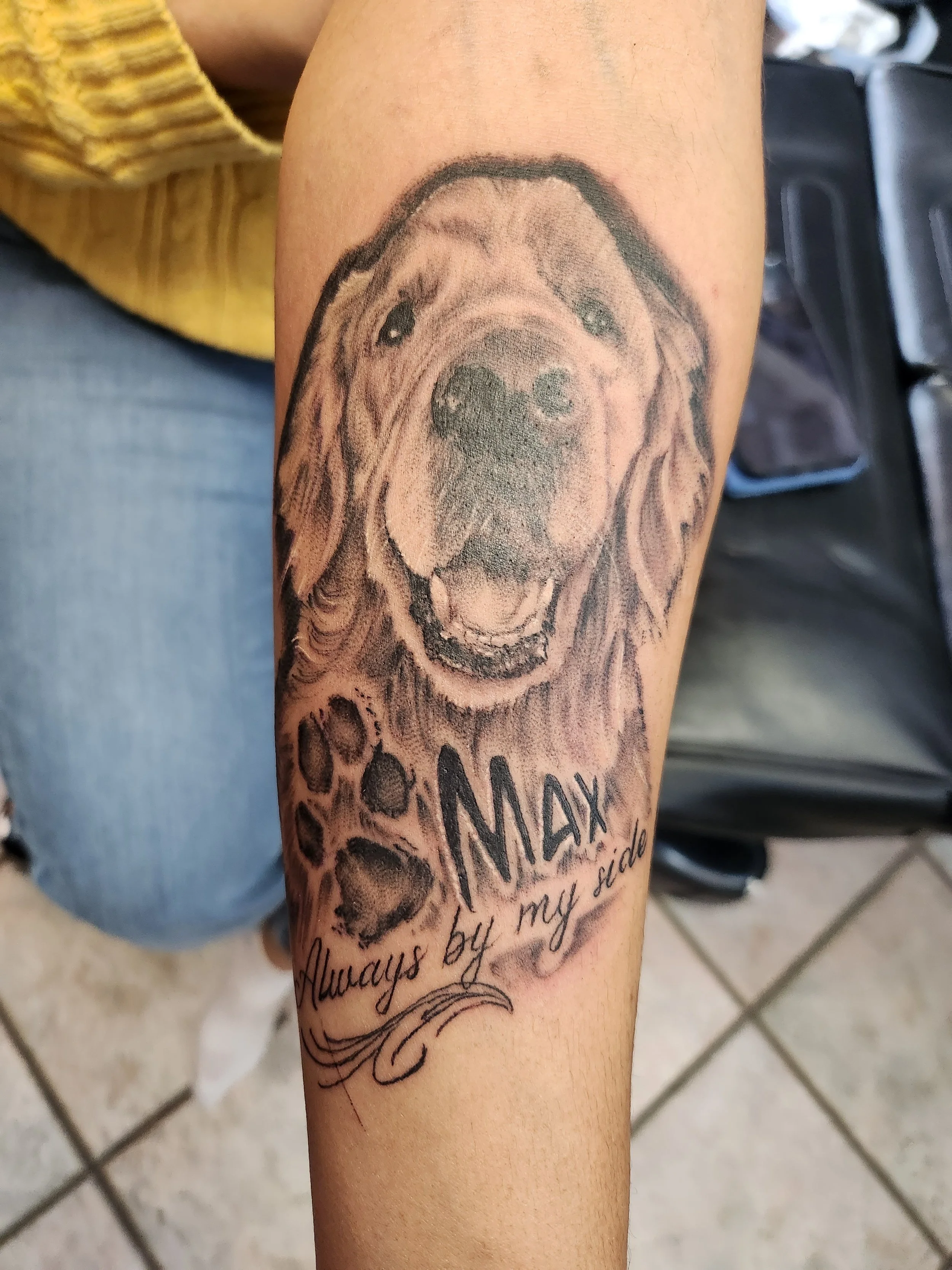 Tattoo of a happy dog, likely a retriever, with the words "MAMA Always by my side" written below, on a person's arm.