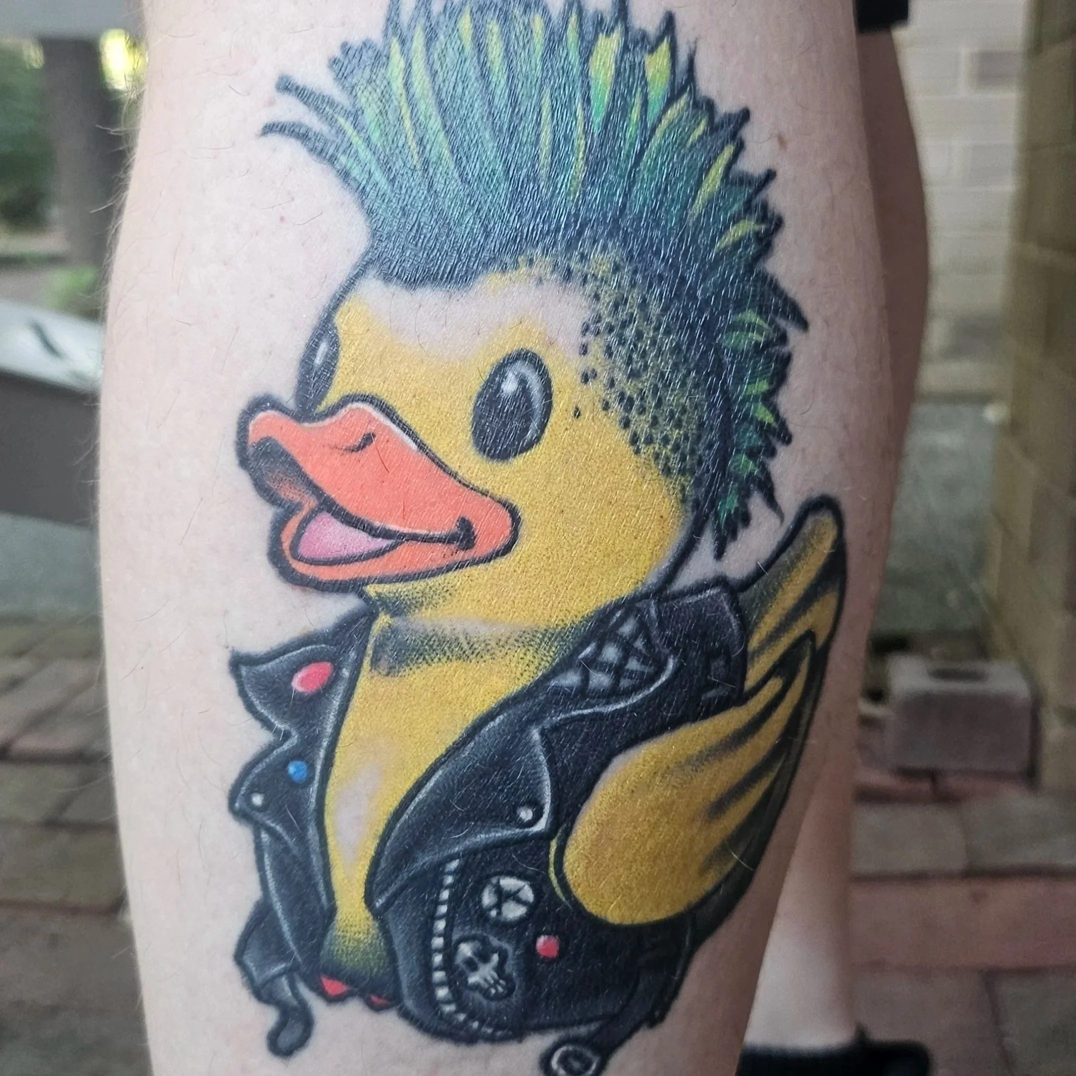 Tattoo of a cartoon duck wearing a leather jacket, with a mohawk hairstyle, holding a video game controller.