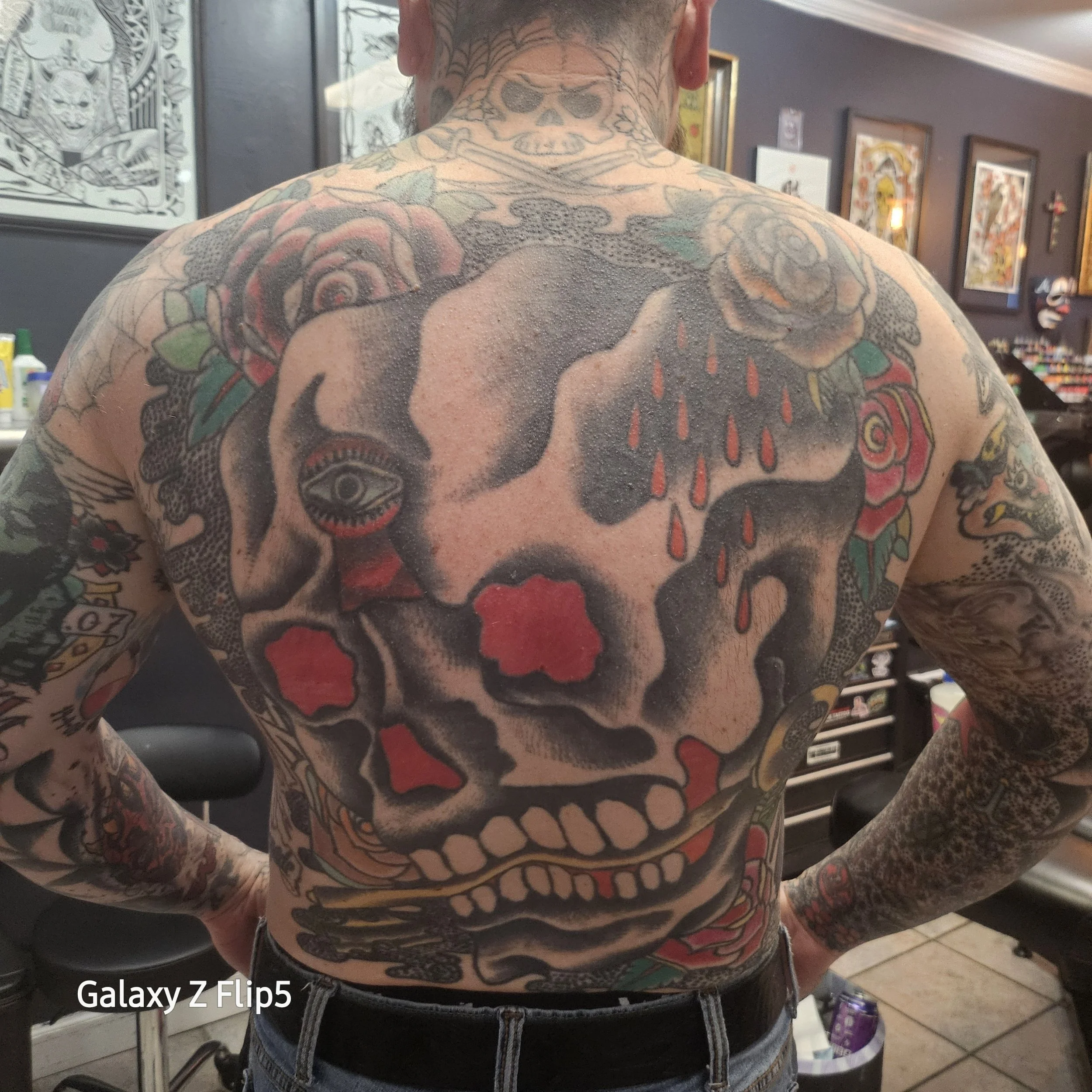 A person with extensive tattoos on their chest and arms, featuring a large tattoo of a skull with red accents, roses, and other designs, in a tattoo studio with framed artwork on the walls.