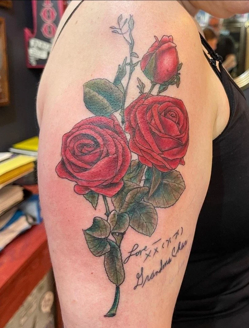 Tattoo of two red roses with green leaves and a stem on a person's upper arm, with a handwritten message that reads "for X, X X) Grandmother