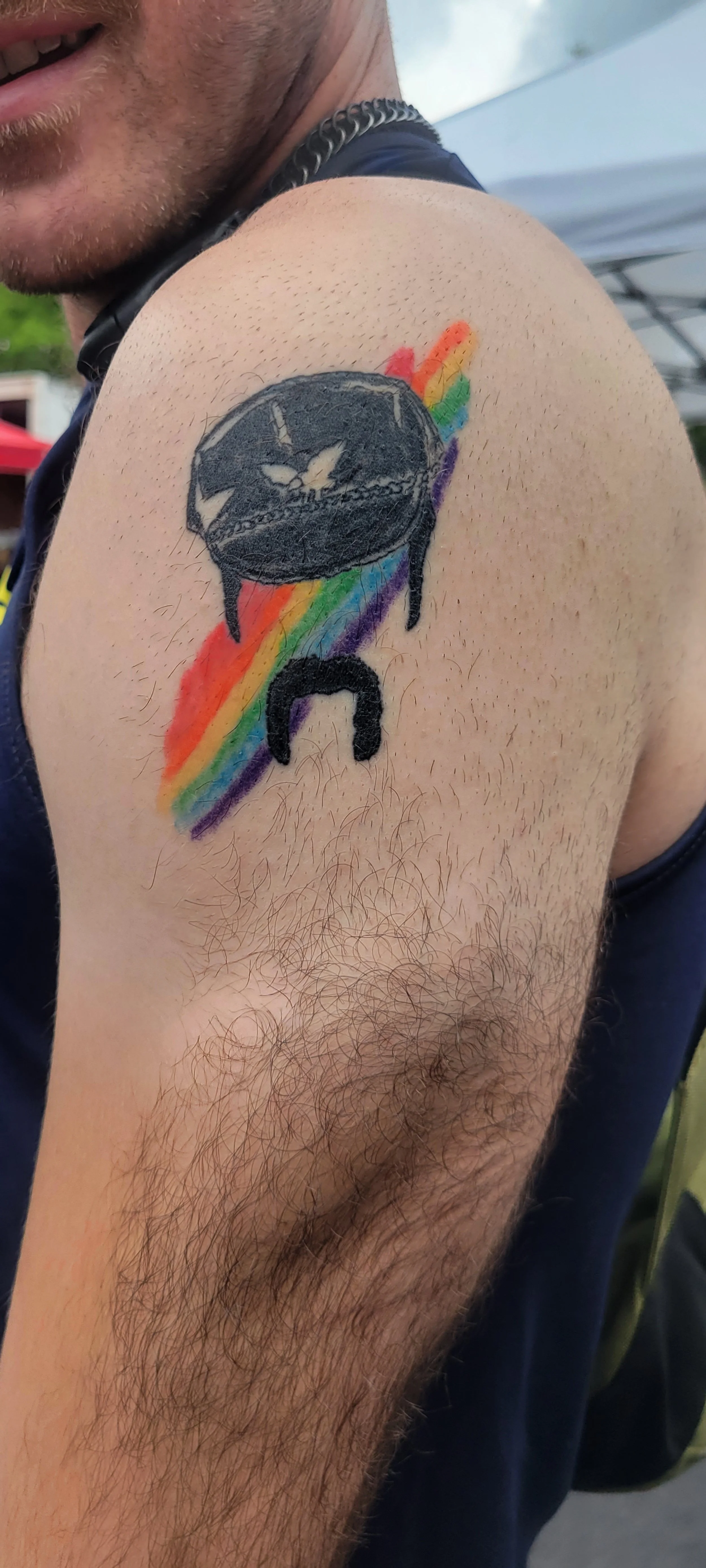 Tattoo of a clock with the hands showing 11:45, dripping black paint, and a rainbow-colored trail behind it on a man's upper arm.