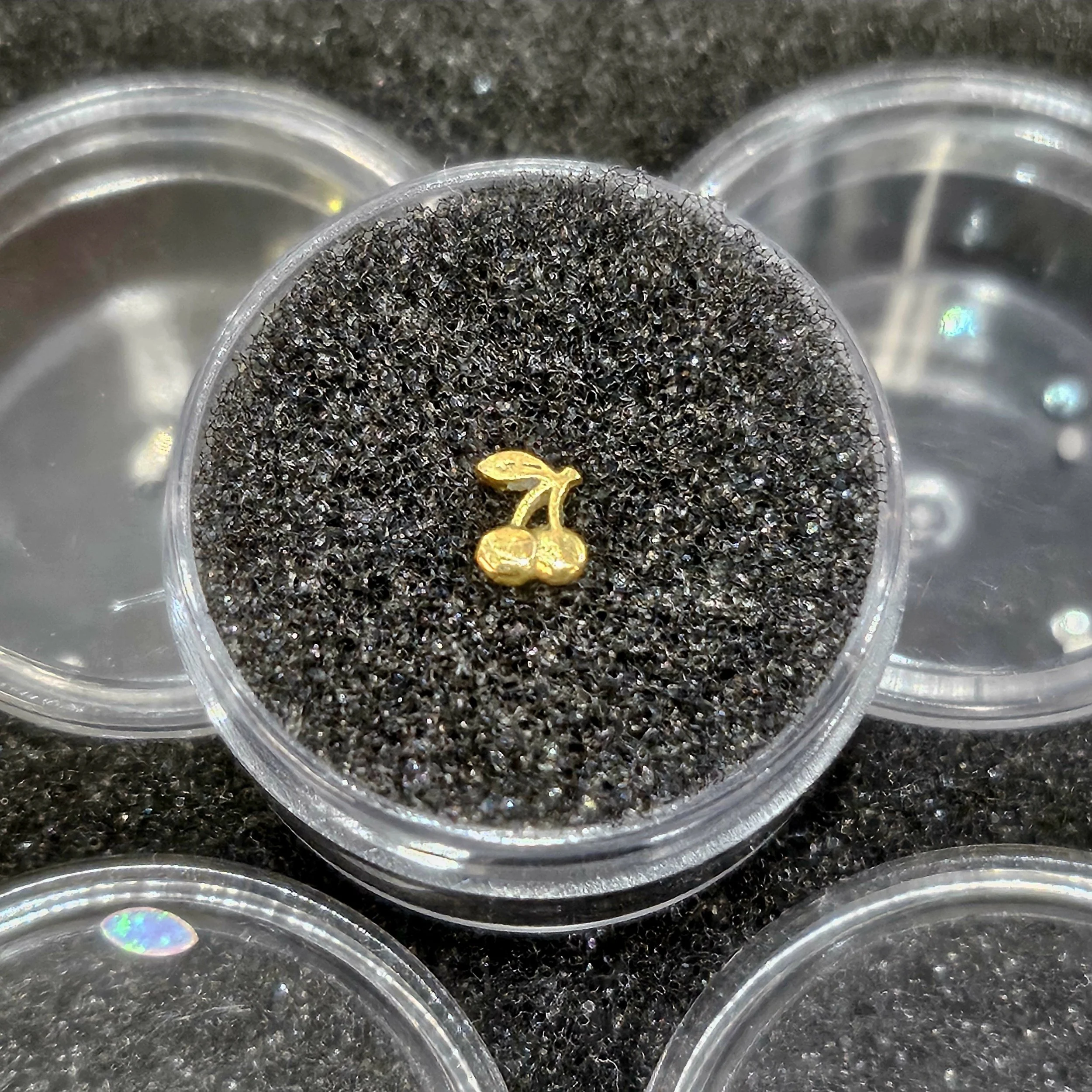 Small gold cherry-shaped jewelry piece inside a transparent container with black foam padding, surrounded by other similar containers.