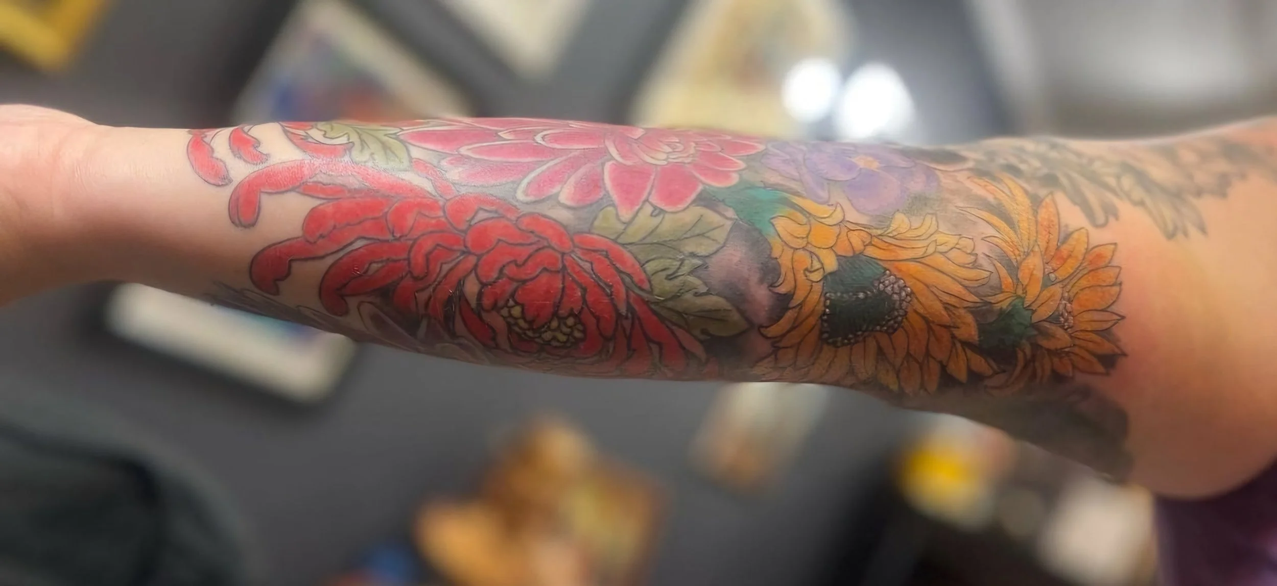 Close-up of a tattooed arm with floral designs, including red, purple, yellow, and orange flowers, on a person's forearm.