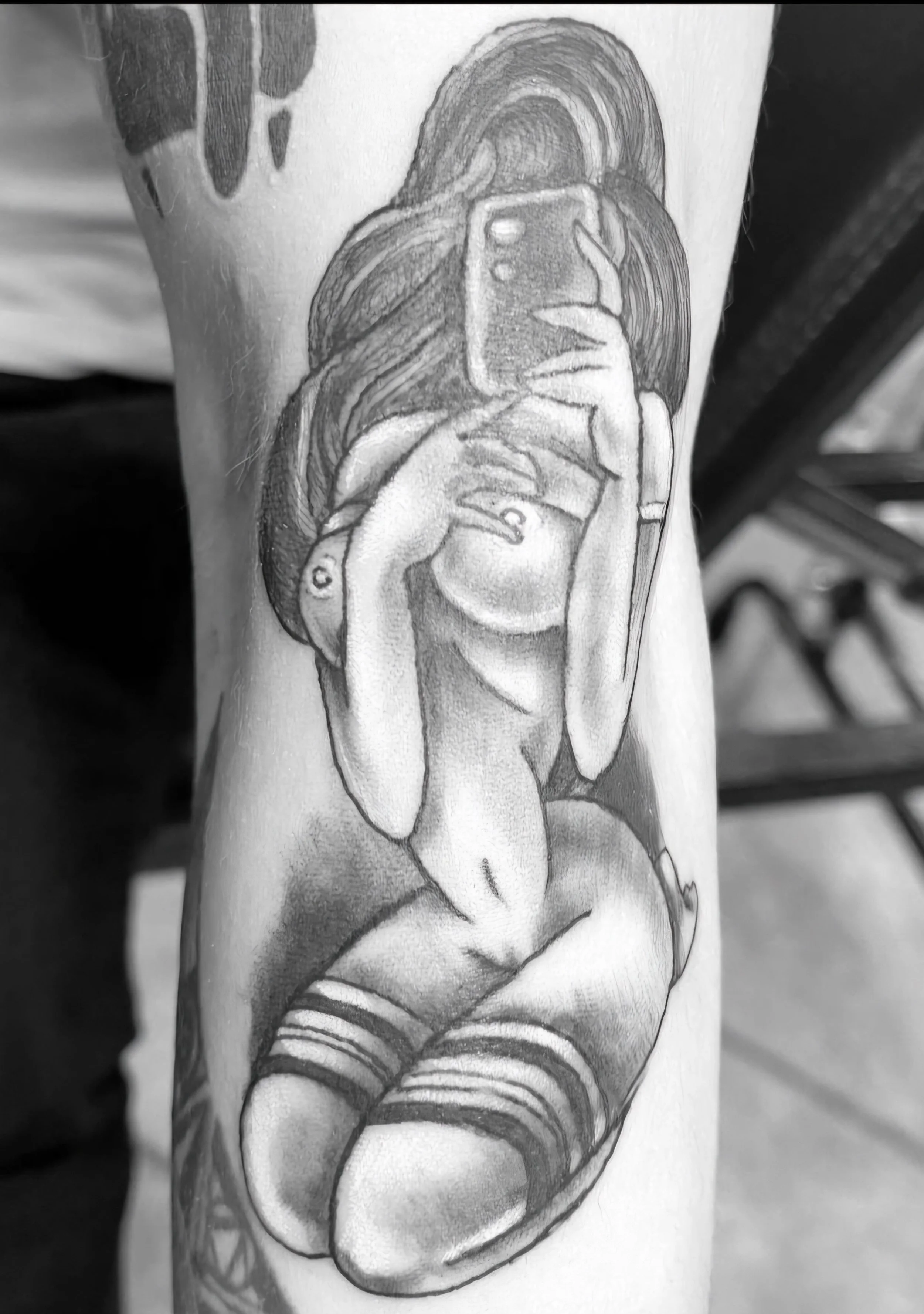 Black and gray tattoo of a nude woman with long hair taking a selfie with a camera phone, sitting with knees up, wearing striped thigh-high stockings.