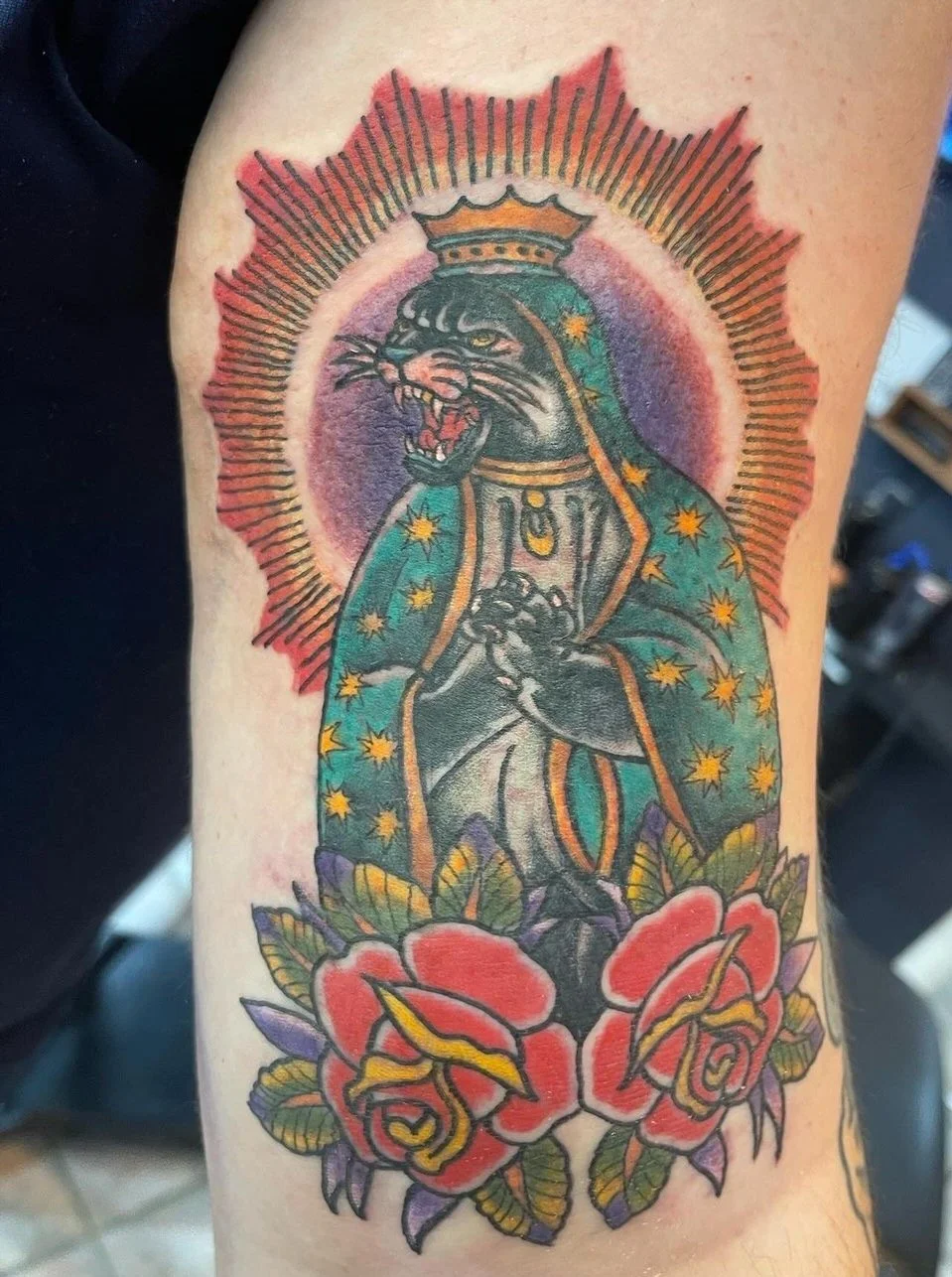 Tattoo of a black panther wearing a crown and a starry cape, with a radiant halo and roses at the bottom.