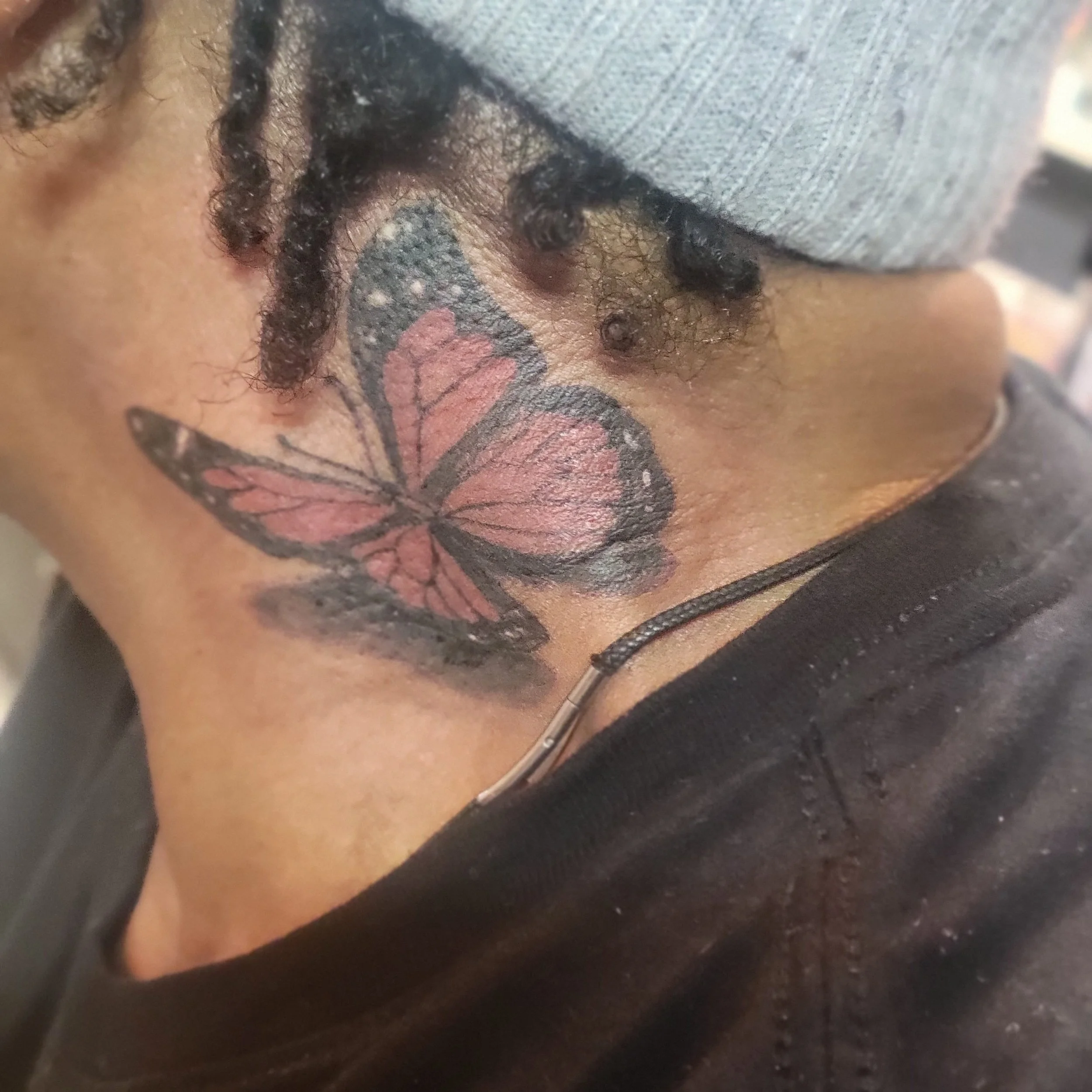 Close-up of a butterfly tattoo on a person's neck, showing pink and black colors, with some hair and a gray cap partially visible.