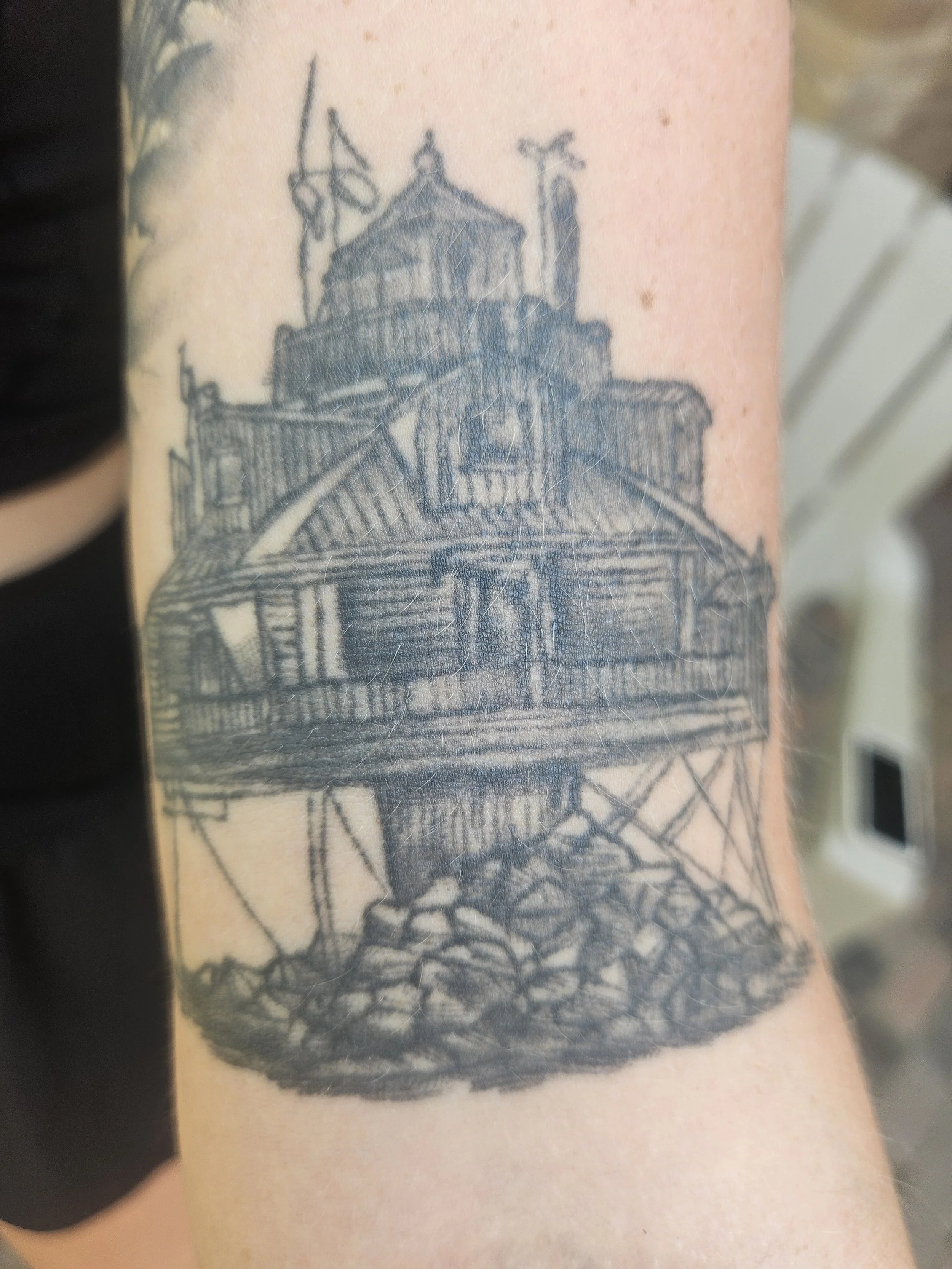 Black and gray tattoo of a house on a rocky foundation, with a staircase leading up to the entrance, surrounded by flagpoles with flags.