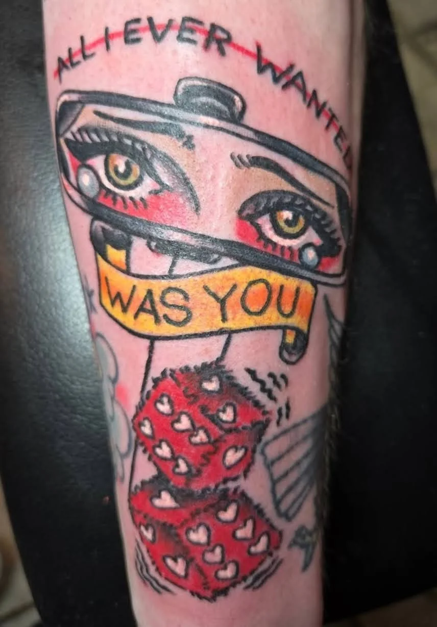 Tattoo of a pair of eyes with makeup, a banner reading 'WAS YOU,' red hearts, and a pair of dice with heart patterns.
