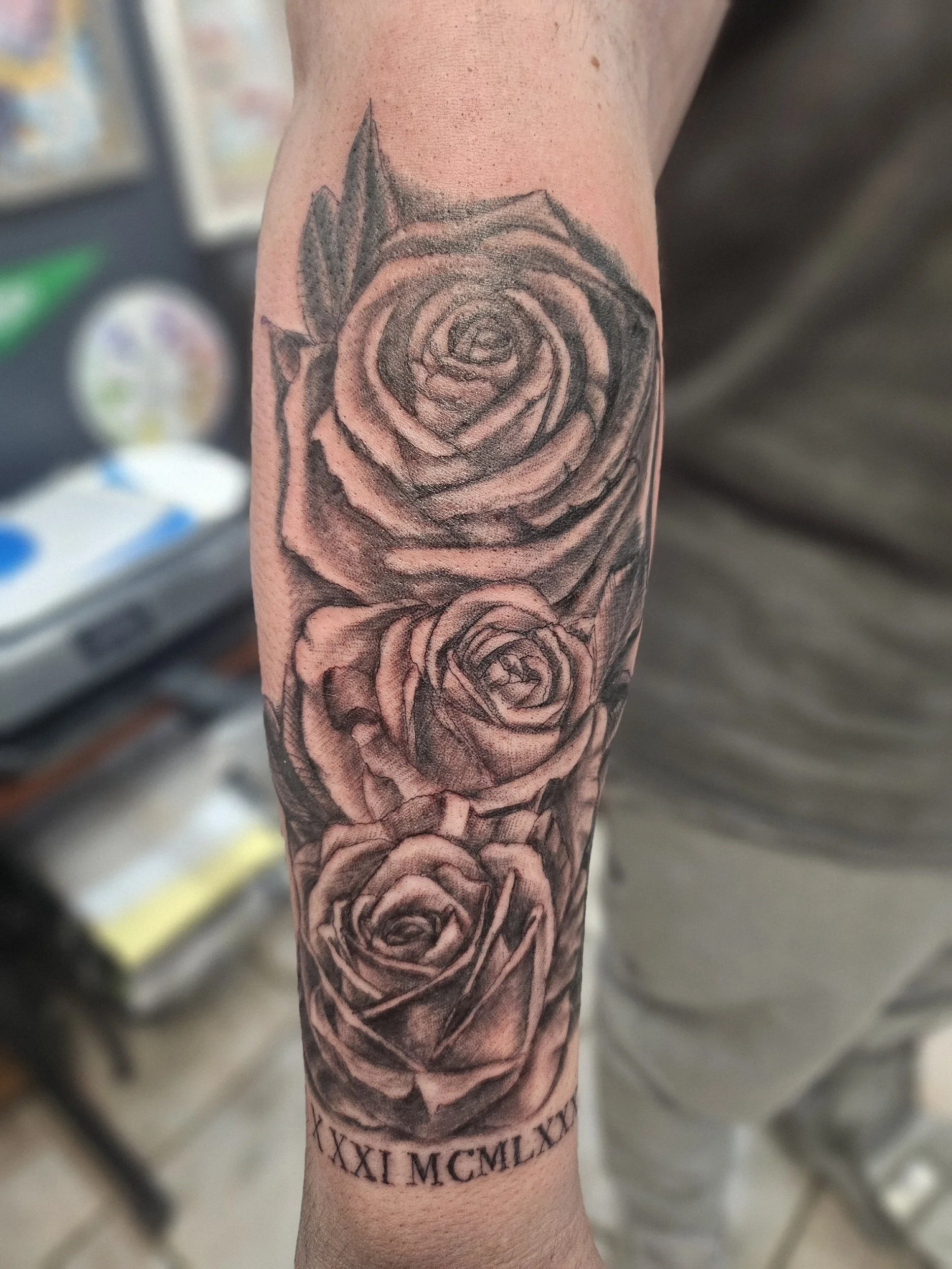 A detailed black and gray tattoo of three roses on a person's forearm, with Roman numerals at the bottom.