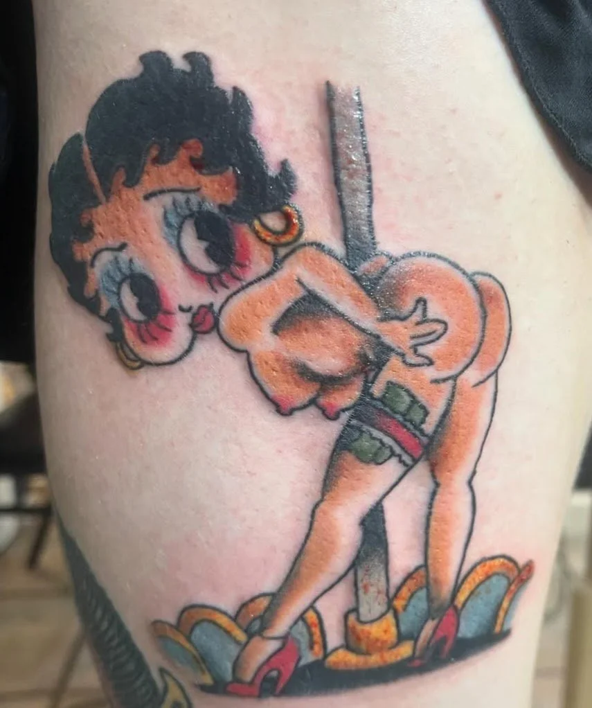 Tattoo of Betty Boop with her lower body as a pin-up girl, wearing red high heels, shorts, and a tank top, leaning forward on a pole.