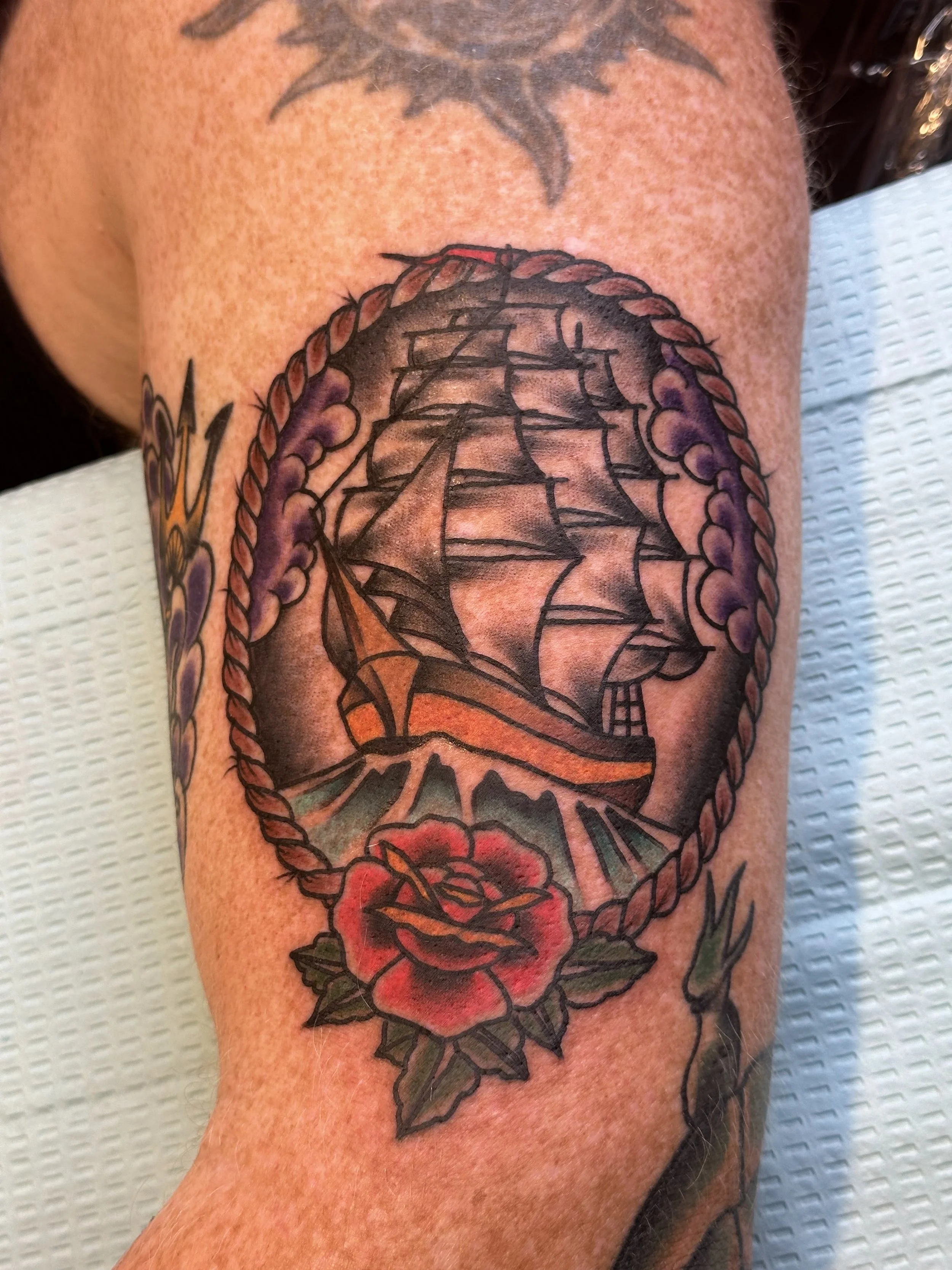 Tattoo of a sailing ship within a rope oval and a red rose at the bottom on a person's arm.