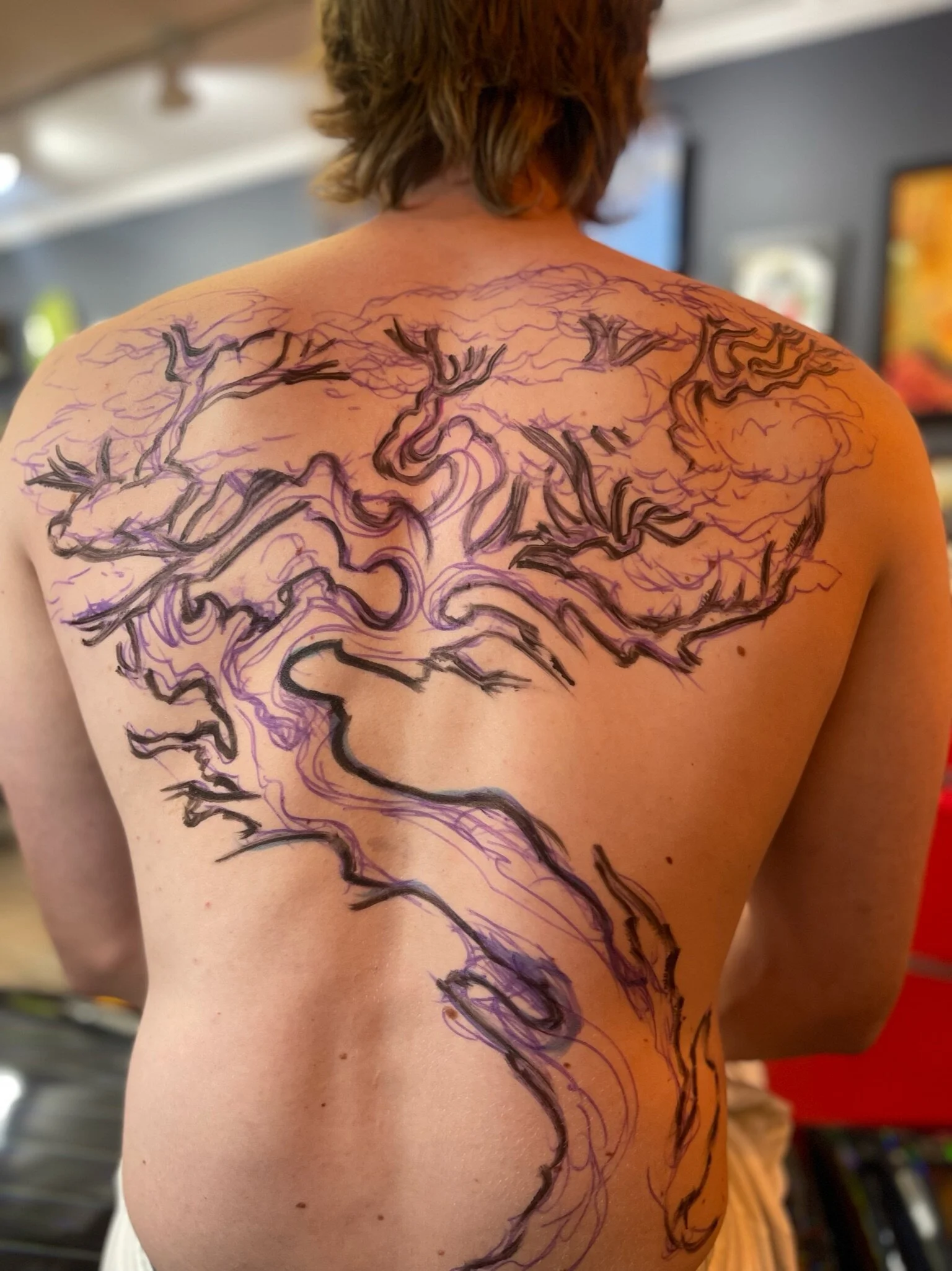 Person with a large back tattoo in progress, with black and purple ink sketches of a tree and its branches covering the upper back and shoulder blades.