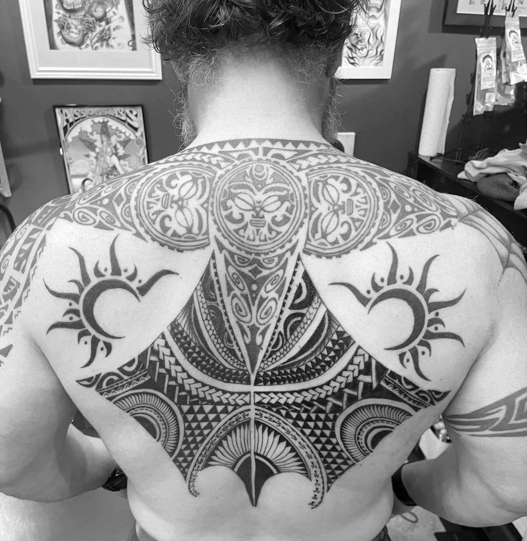 Back view of a person with a large intricate Polynesian-style tattoo covering the shoulders and upper back, including sun or moon symbols and detailed geometric patterns.