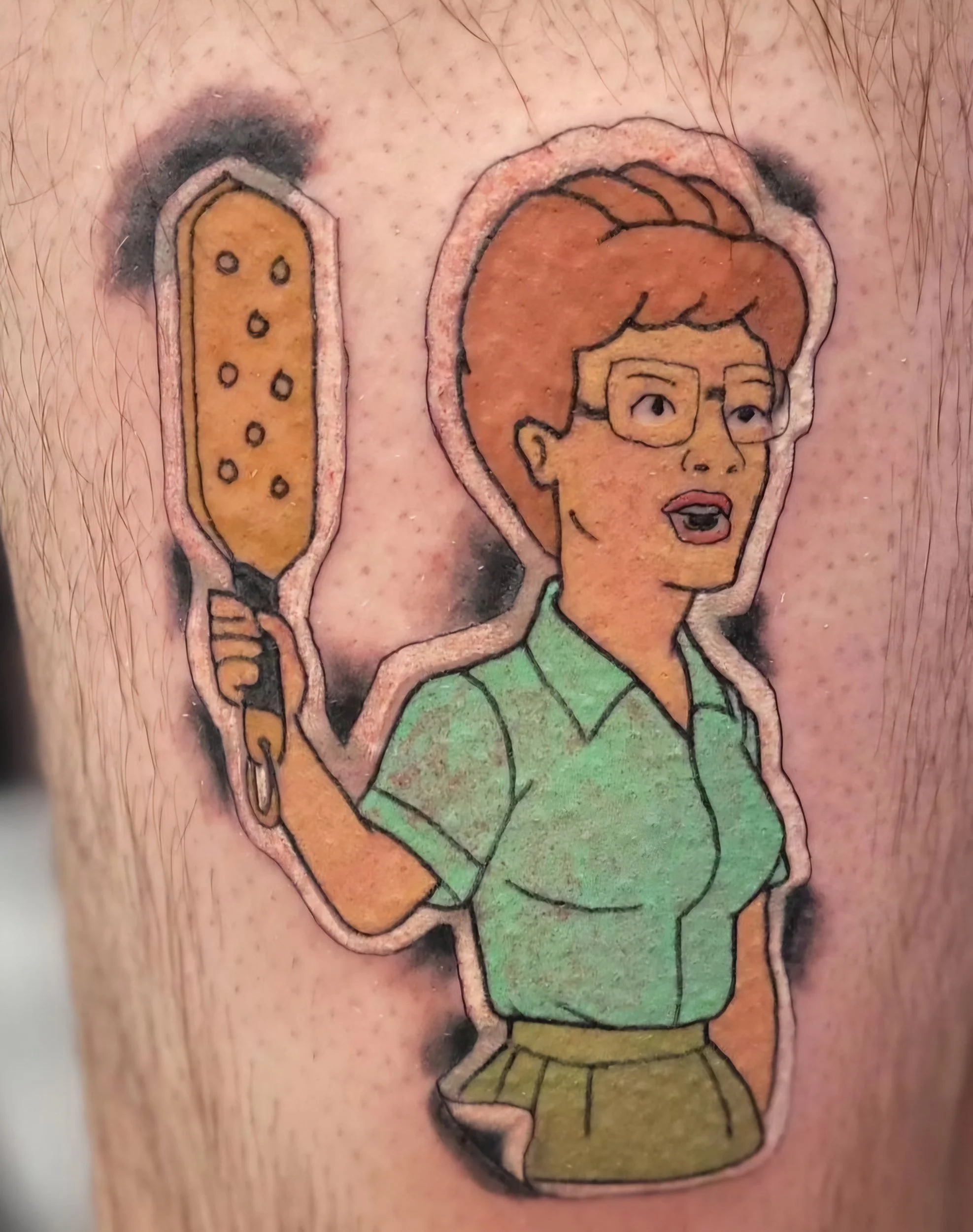 Tattoo of a cartoon woman with glasses, holding a spatula, wearing a green shirt and brown pants.
