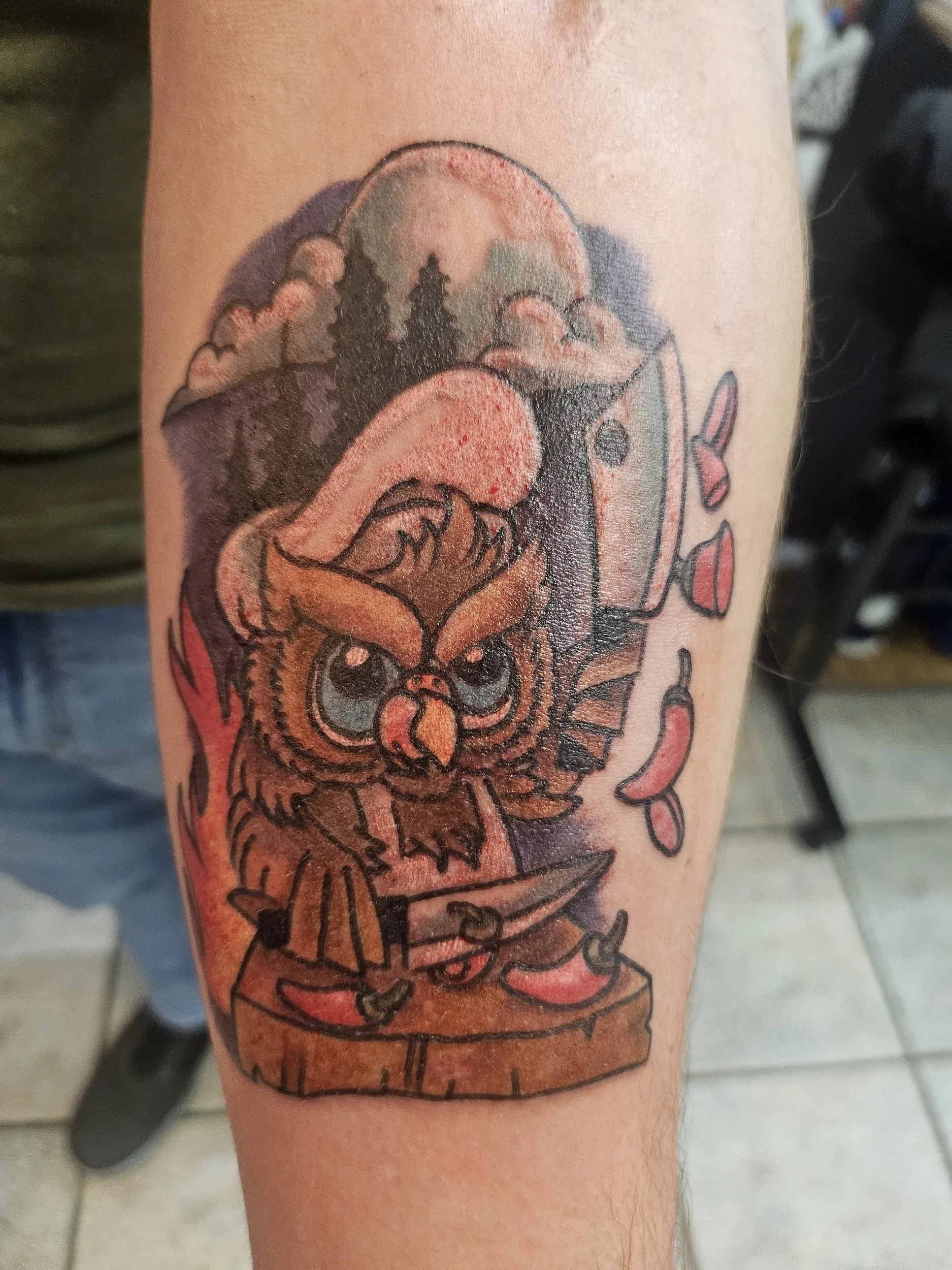 Tattoo of an owl with large eyes, fiery red flames, a background of forest trees, a knife on a wooden surface, and chili peppers around it.