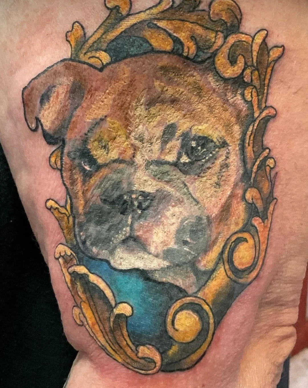 Tattoo of a dog's face surrounded by ornate gold and blue decorative elements.
