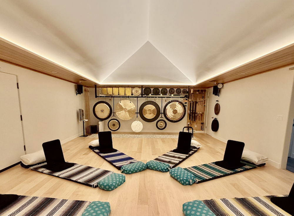 Photograph of Blosm Studio space where breathwork sessions occur. White room with central peaked ceiling, floor mats and pillows, and the back wall has hanging gongs and chimes of different sizes.