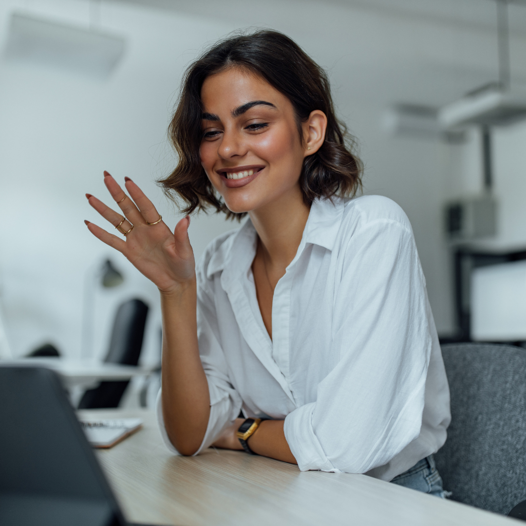 A young woman with dark wavy hair smiling and waving while looking at her laptop in an office setting.