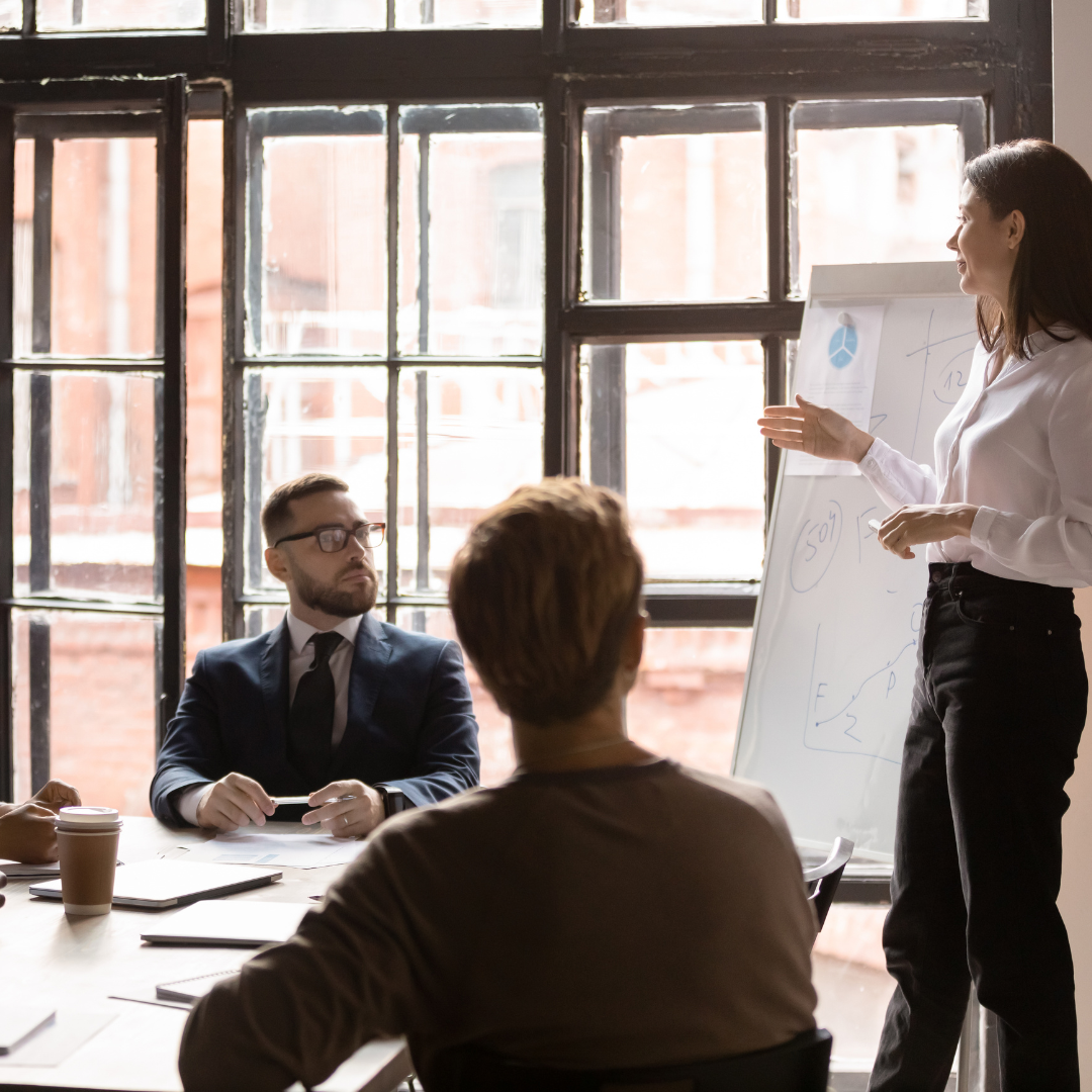 A woman giving a presentation in a meeting room with three people seated around a table, large windows in the background letting in natural light.