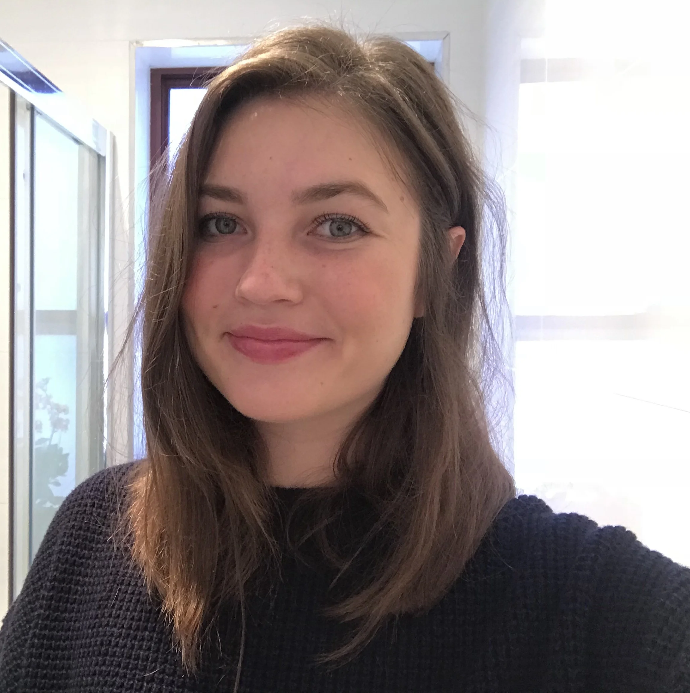 A young woman with shoulder-length brown hair, blue eyes, and a slight smile, wearing a black textured top, standing indoors in front of a window with sunlight streaming in.
