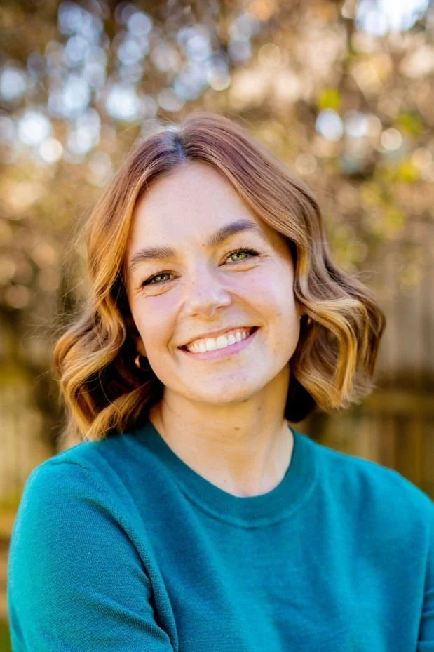 A young woman with shoulder-length wavy red hair, smiling in an outdoor setting with blurred trees and sunlight in the background, wearing a teal long-sleeve shirt.