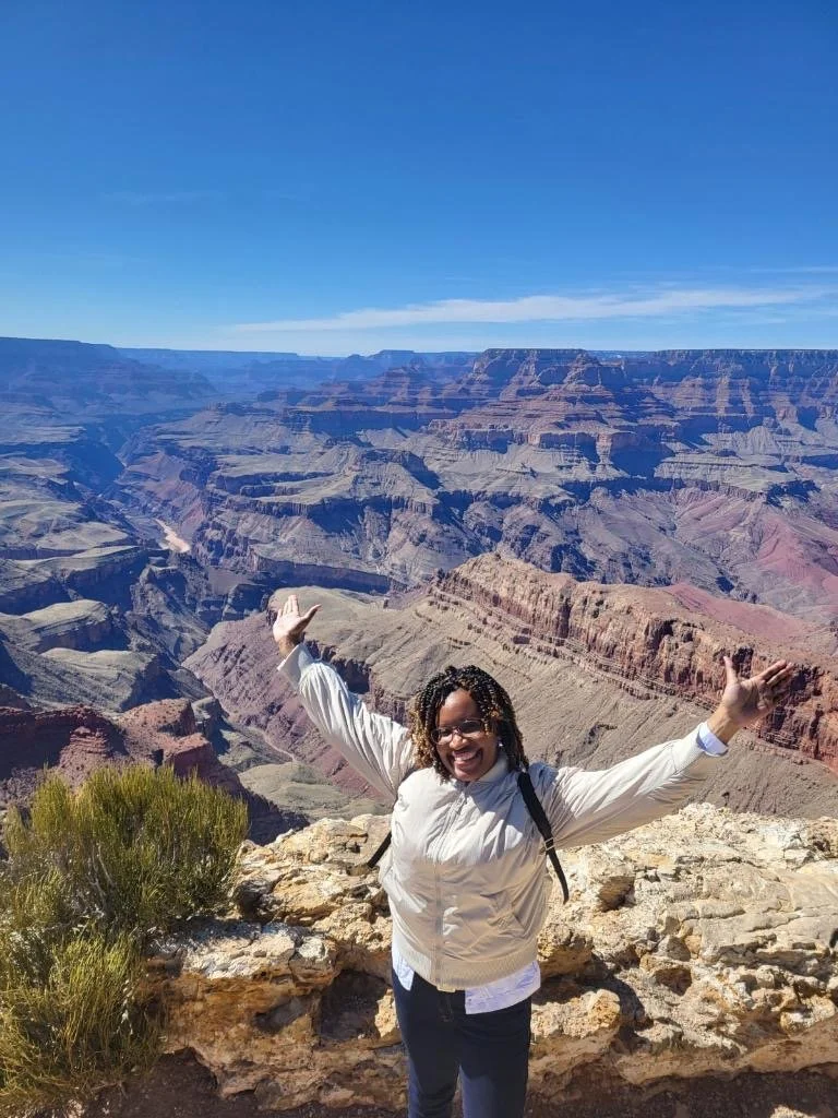 A woman standing at the Grand Canyon with arms outstretched, smiling, wearing a light-colored jacket and sunglasses, with a blue sky and canyon in the background.