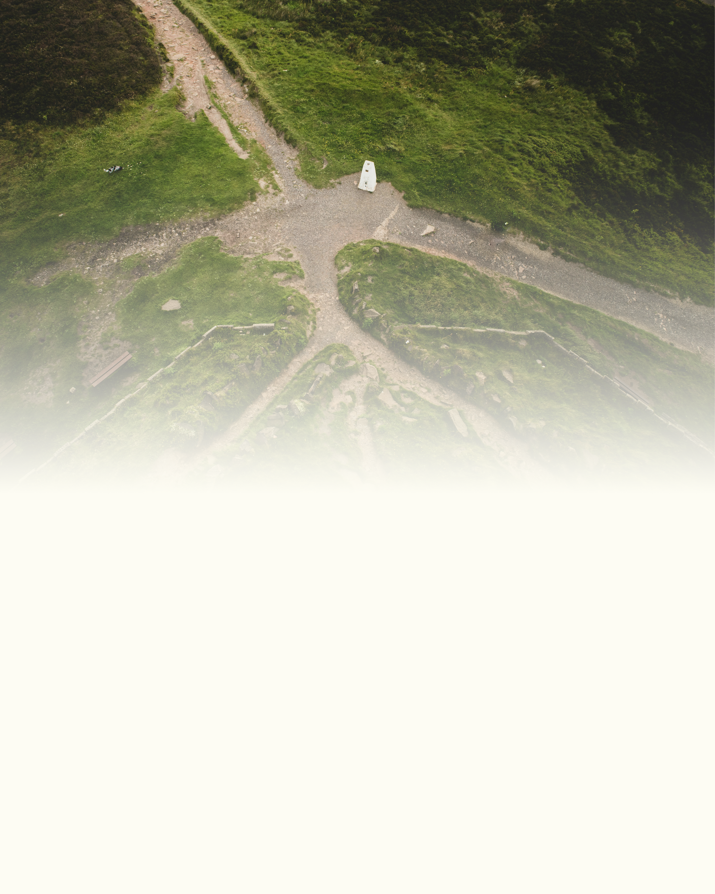 Aerial view of a circular hiking trail intersecting with a gravel path near a green hillside, with a small monument or marker in the center.