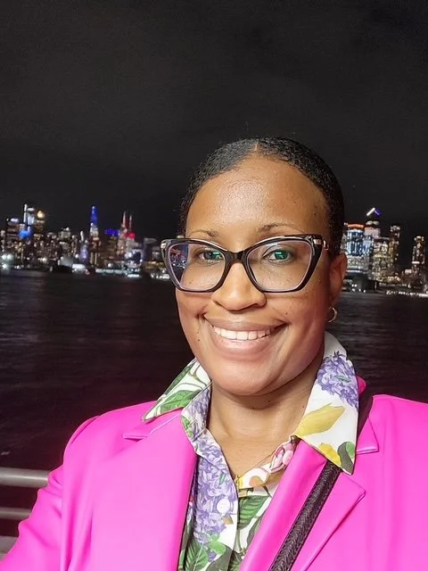A woman smiling at night with a city skyline in the background, wearing glasses, a pink blazer, and a floral shirt.