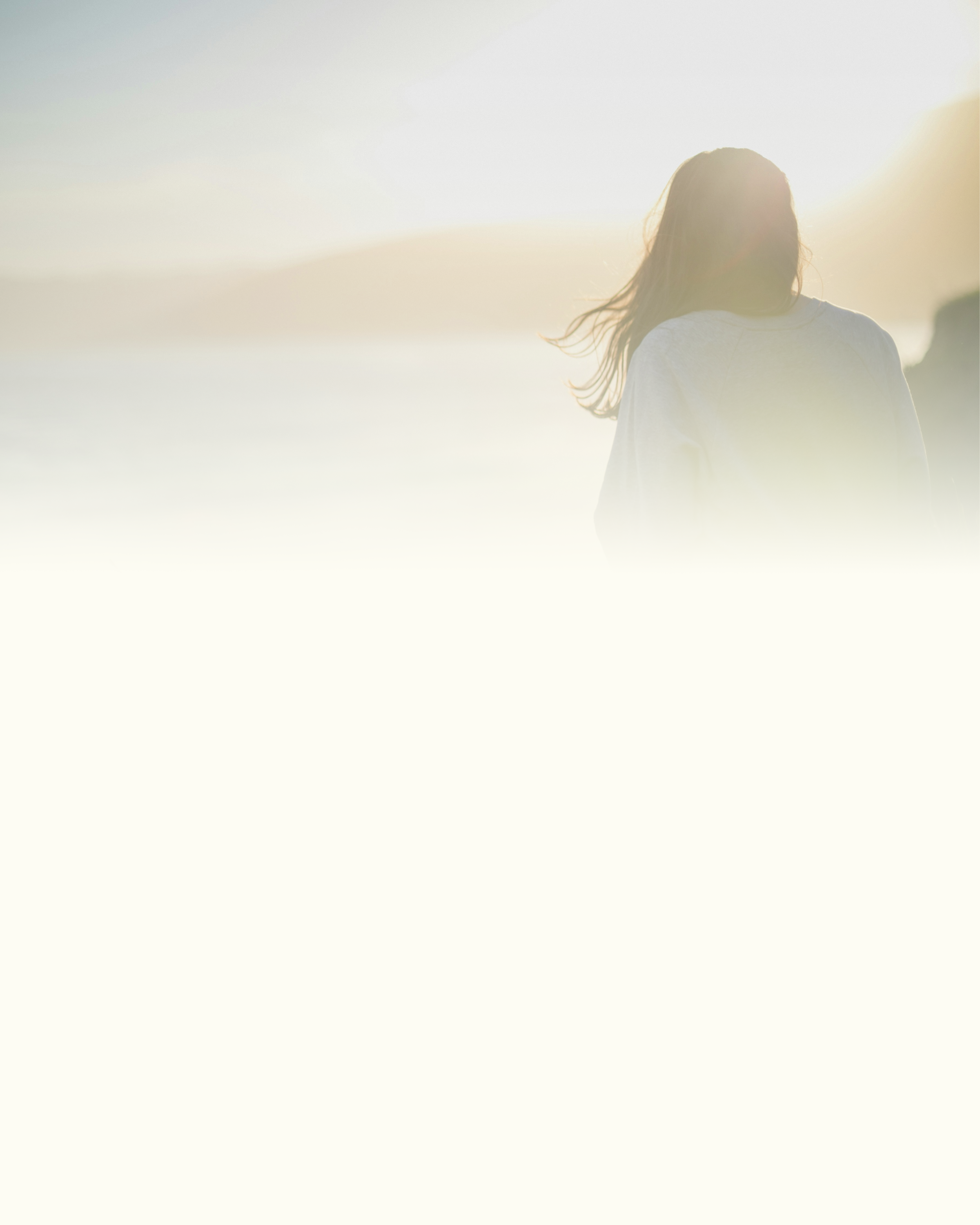 A person with long hair standing on a beach during sunset or sunrise, with bright sunlight creating a silhouette.