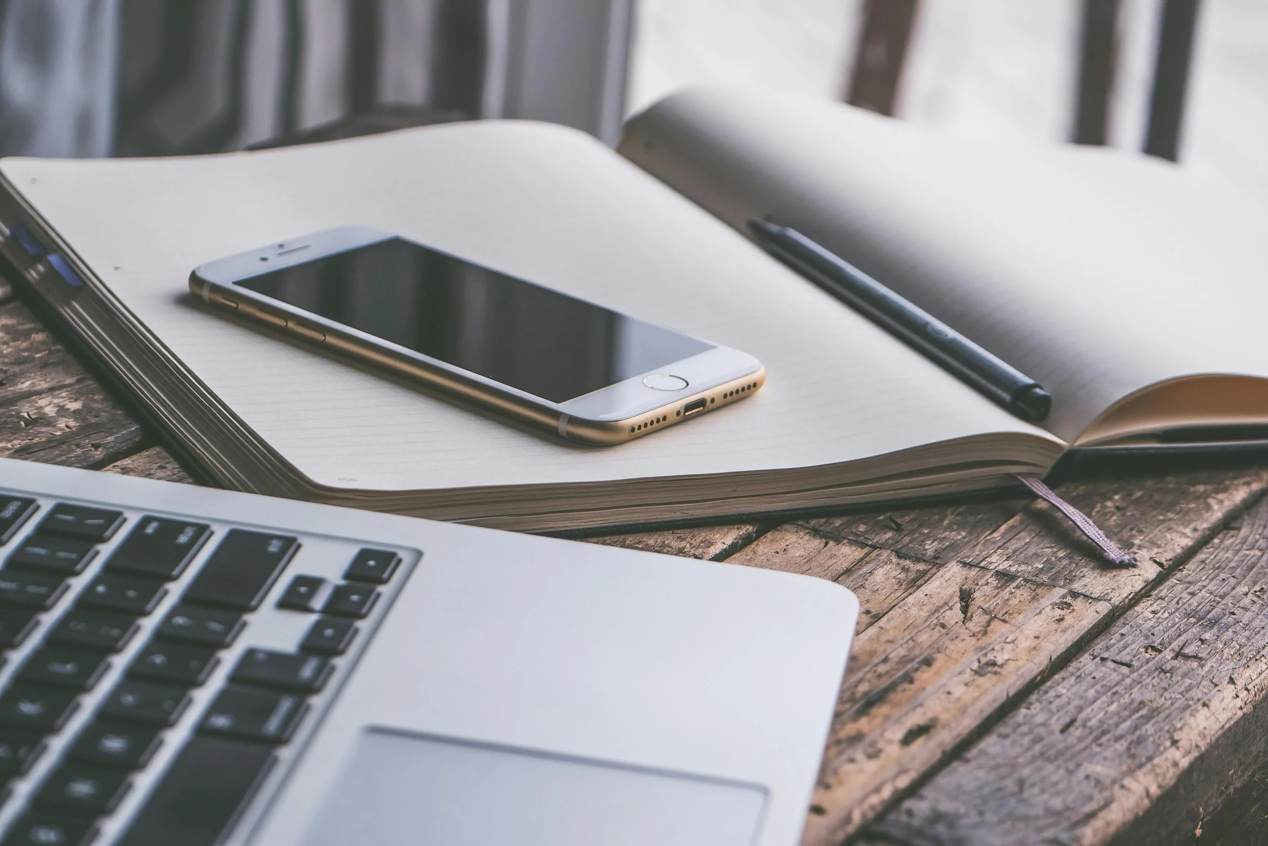 Open notebook with a pen, a smartphone, and a laptop on a rustic wooden table.