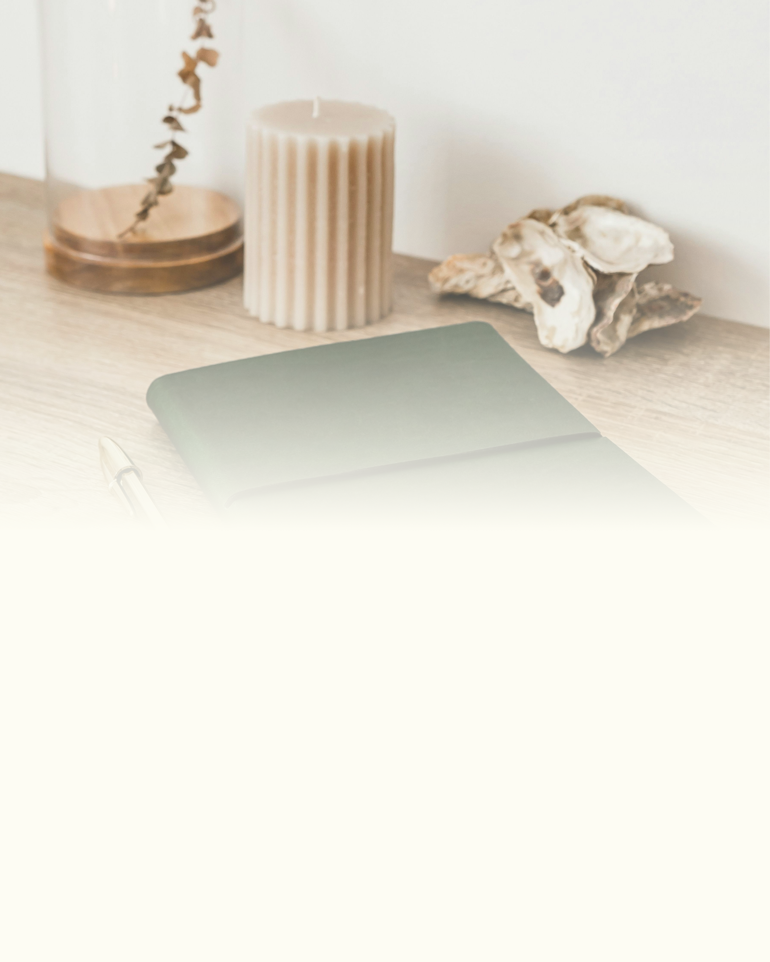 A wooden desk with a beige ribbed candle, a small branch of dried flowers or leaves, a notebook with a pen, and pieces of driftwood or decorative rocks.