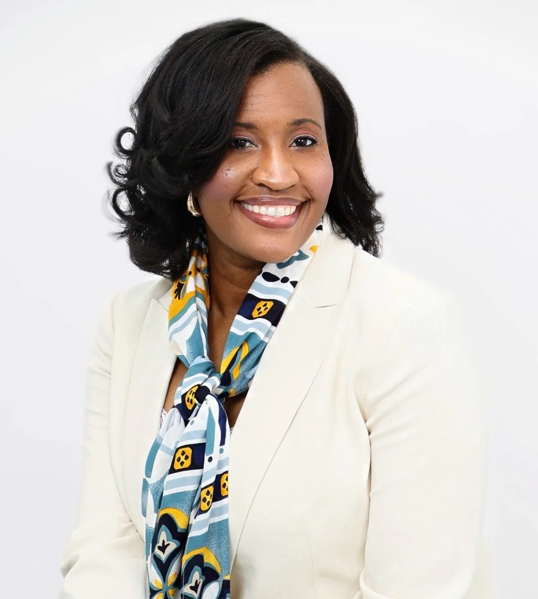 A smiling woman wearing a white blazer and a colorful patterned scarf with black, yellow, and blue details, against a plain white background.