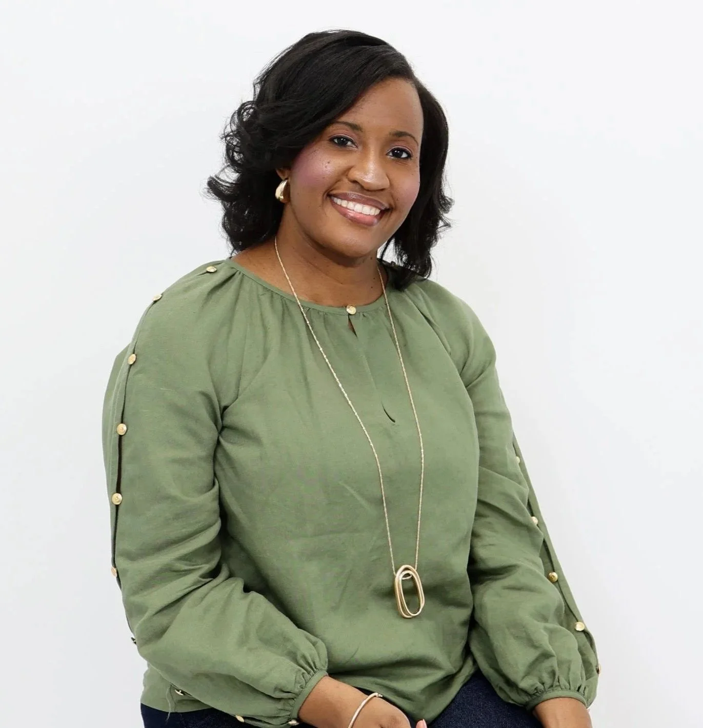 A woman with black hair, smiling, dressed in a green blouse with gold buttons on the sleeves, wearing a long necklace with a gold pendant, posing against a plain white background.