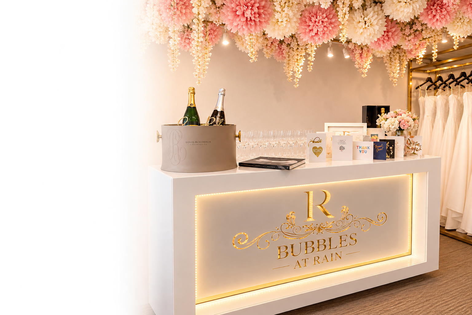 Decorated vanity with pink and cream paper pom-pas and hanging flowers, champagne bottles, empty glasses, thank you cards, and wedding dresses on the side, with a lit sign reading 'Bubbles at Rain'.