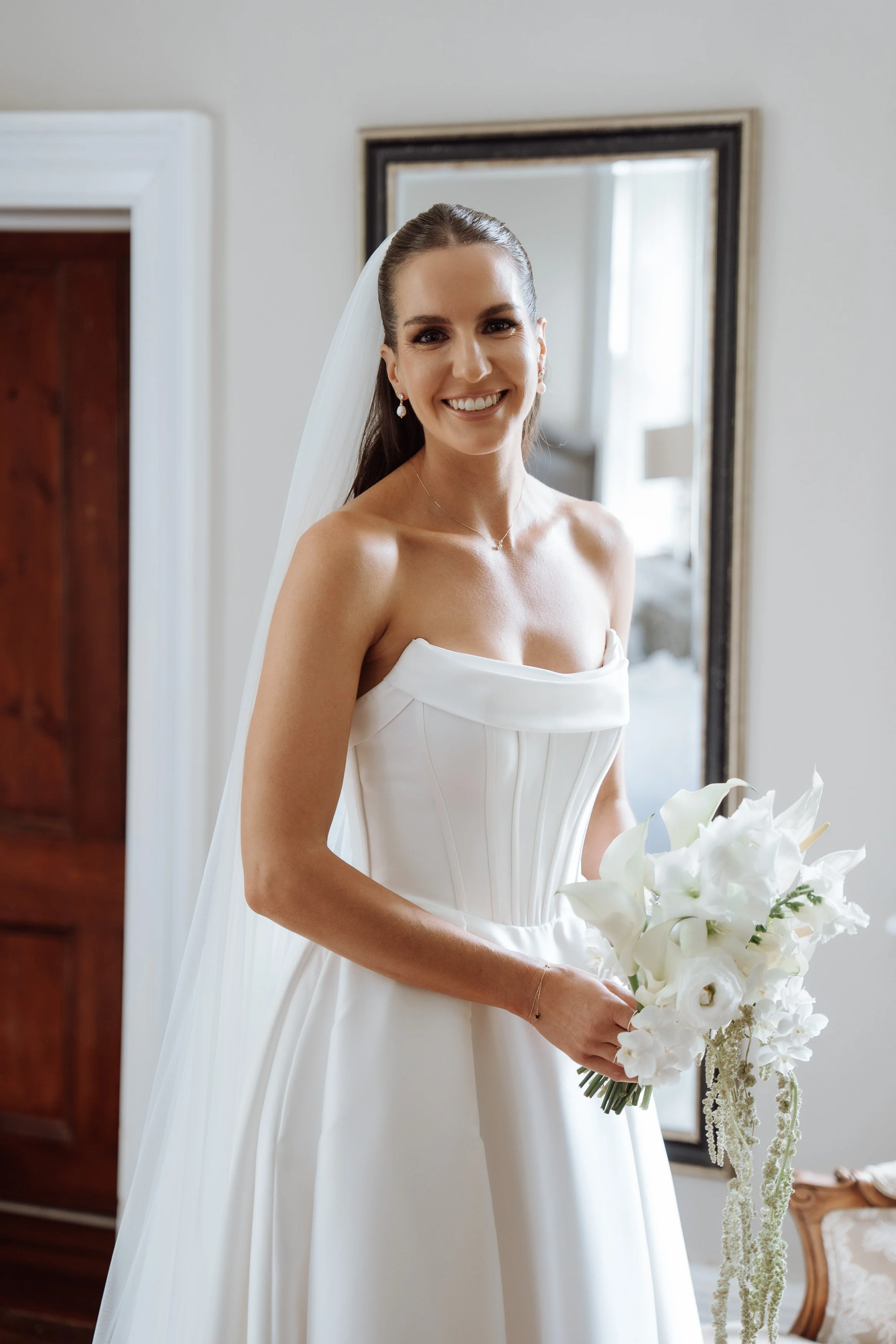 A bride in a strapless white wedding gown holding a bouquet of white flowers and smiling.