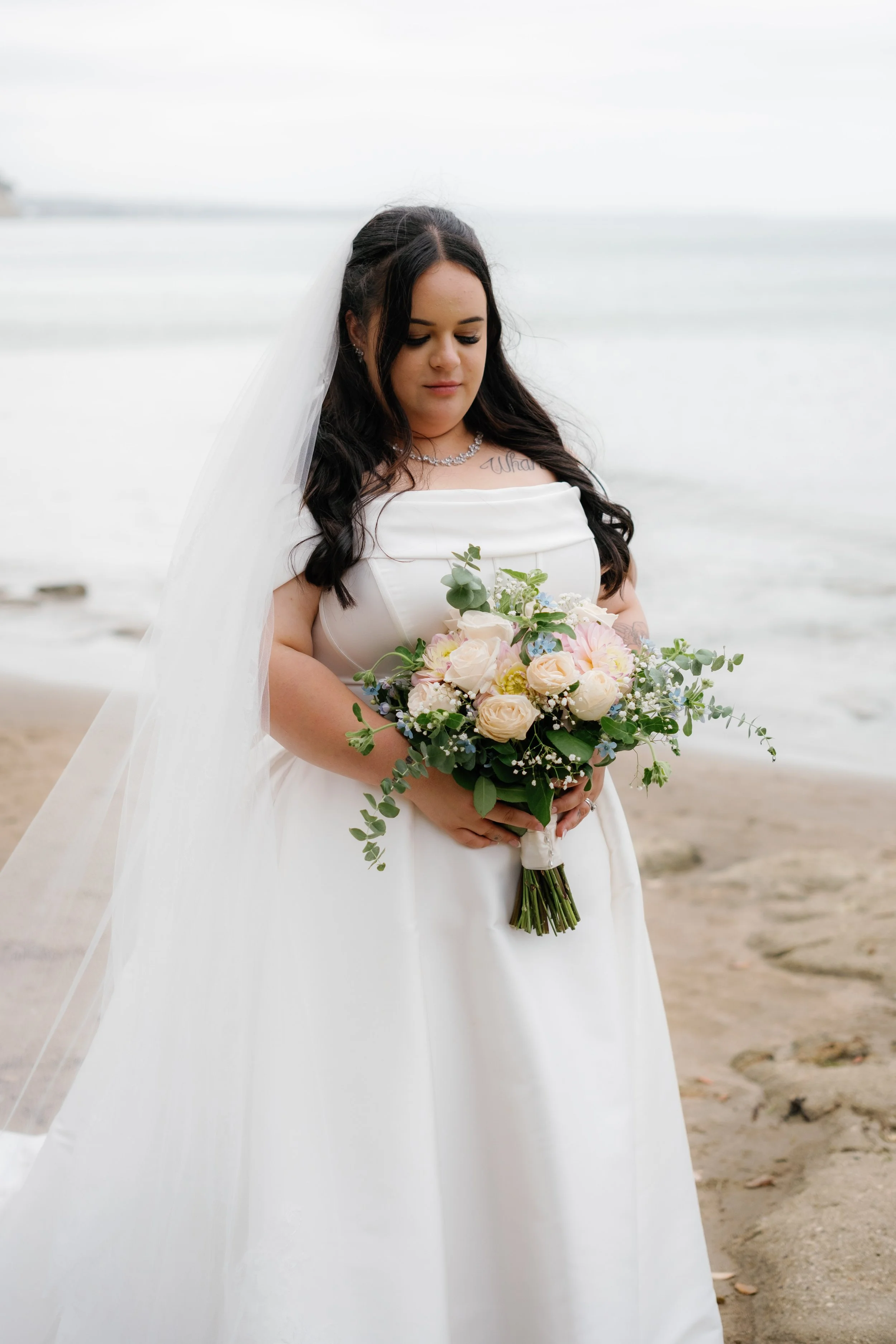 A bride in a white wedding dress holding a bouquet of pink and white flowers at the beach.