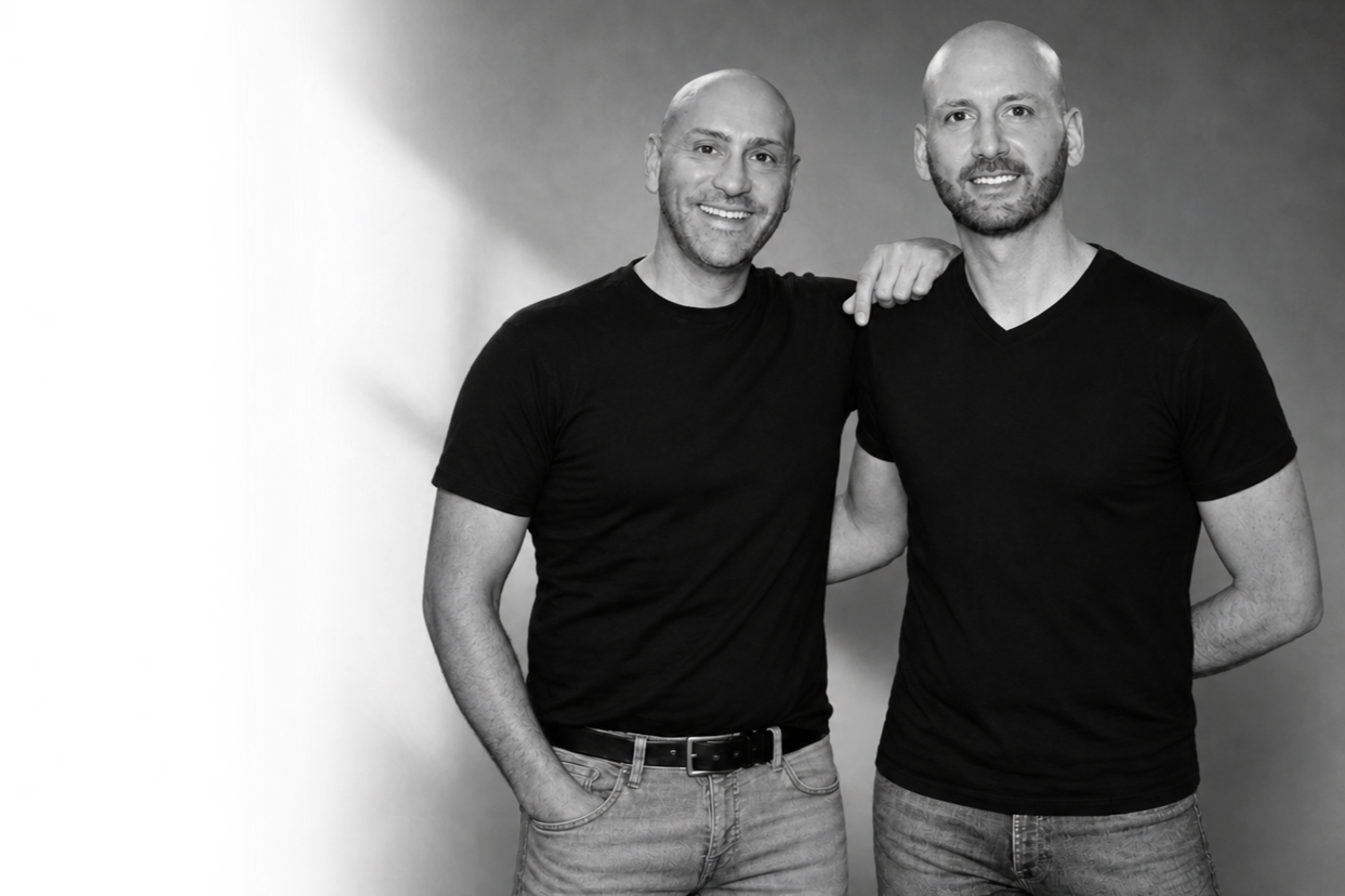 Two men standing together, smiling, with one man's arm around the other's shoulder, wearing black t-shirts and jeans, in front of a plain background.