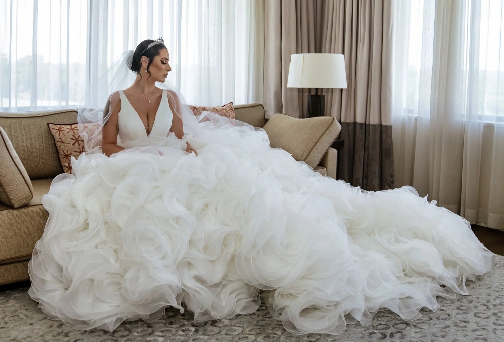 A woman in a wedding dress sitting on a sofa in a room with large windows and beige curtains.