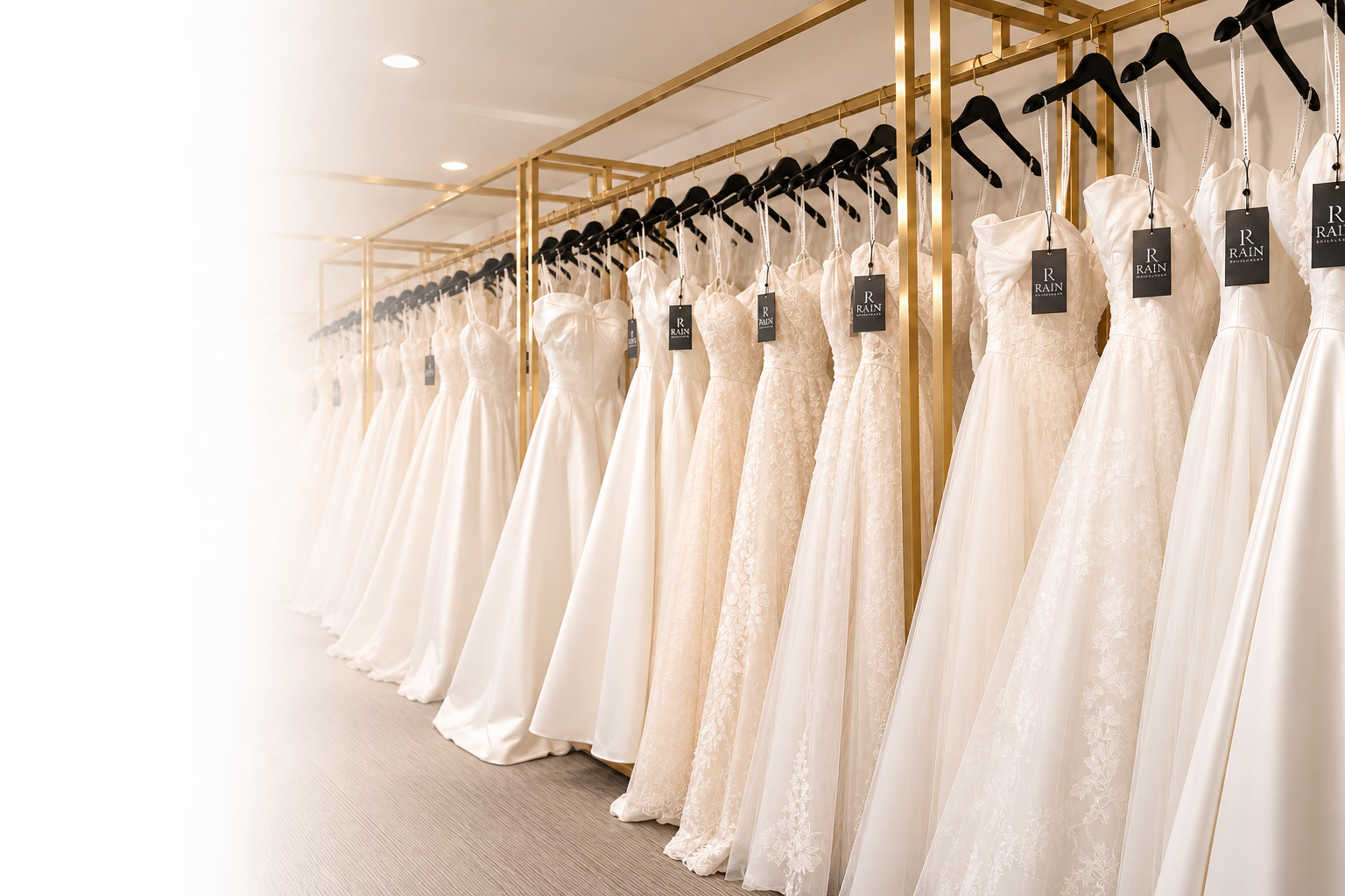 A row of elegant wedding dresses hanging on black hangers with black tags labeled 'R Rain' on a gold clothing rack in a bridal boutique
