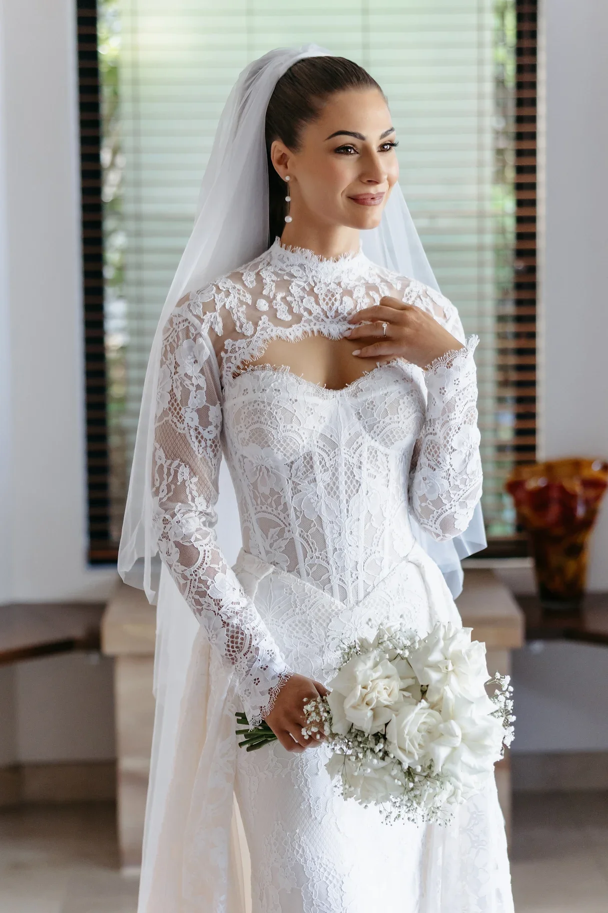 A bride in a white lace wedding dress holding a bouquet of white roses, standing indoors in front of window blinds, smiling softly.