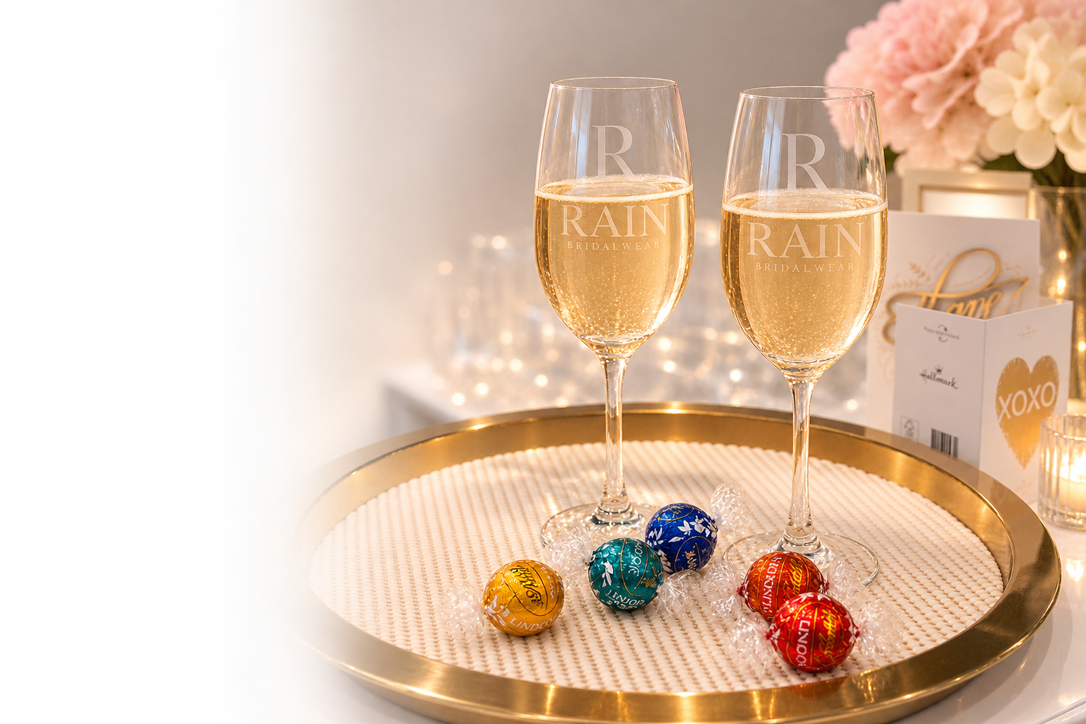 Two champagne glasses filled with bubbly, placed on a gold-rimmed tray with wrapped chocolates. Background includes floral arrangements and greeting cards.