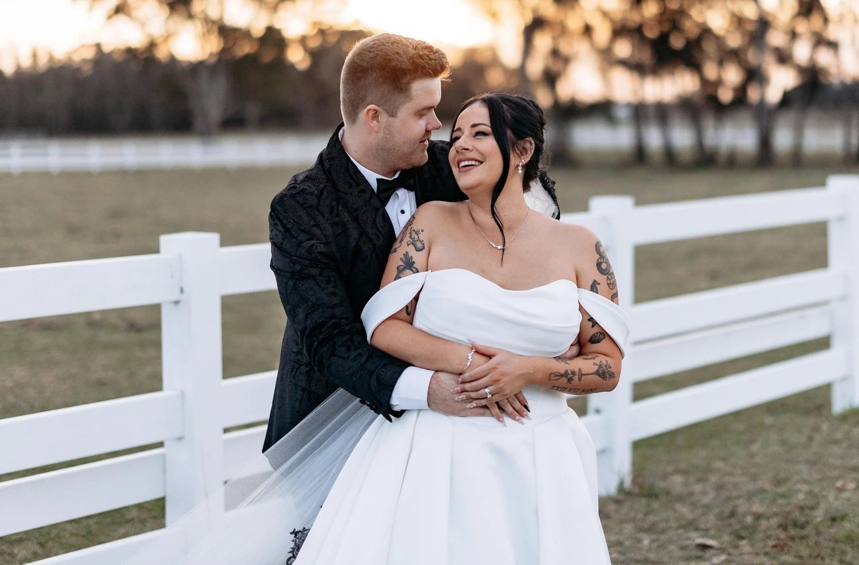 A couple dressed in wedding attire standing outdoors near a white fence, smiling and looking at each other during sunset.