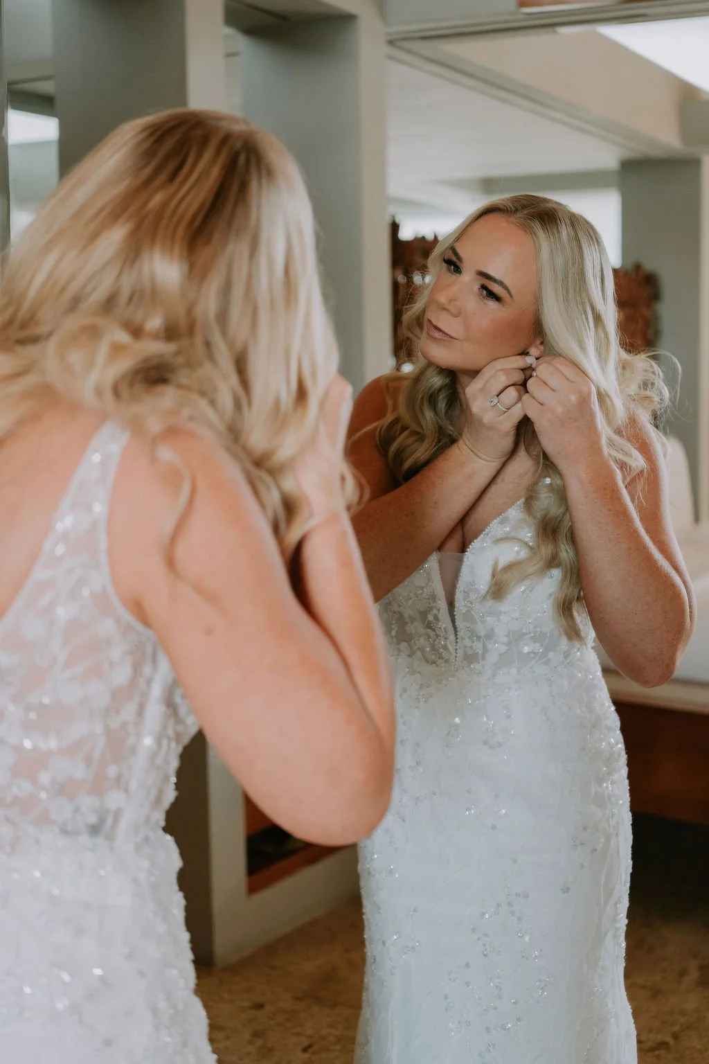 A bride with long blonde hair in a white wedding dress is looking at her reflection in a mirror while adjusting her earring, with another woman helping her.