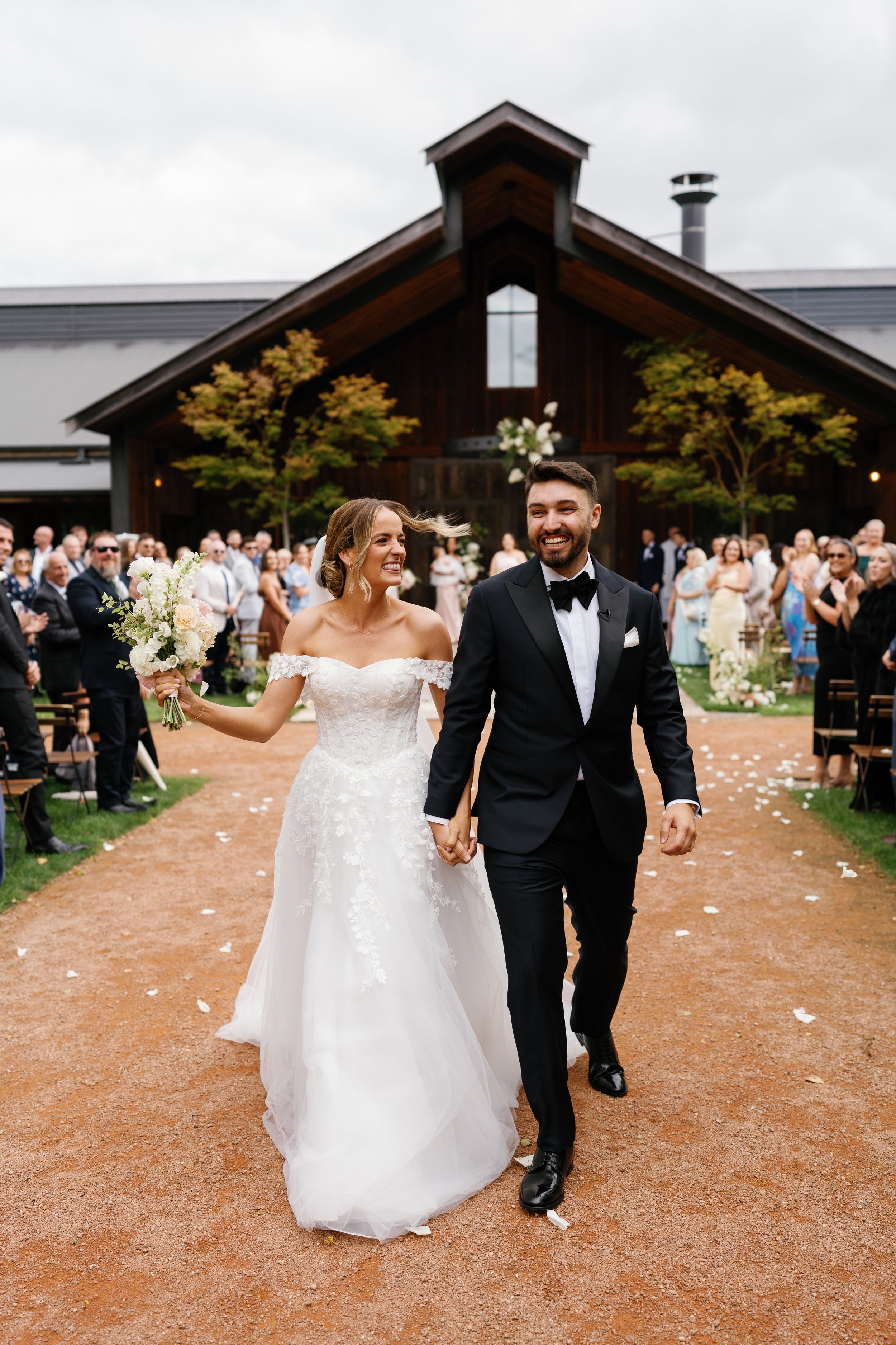 A newlywed couple walking hand in hand down an outdoor aisle, smiling at each other. The bride is wearing a white wedding gown and holding a bouquet, while the groom is in a black tuxedo. Guests are standing on either side, clapping and celebrating a