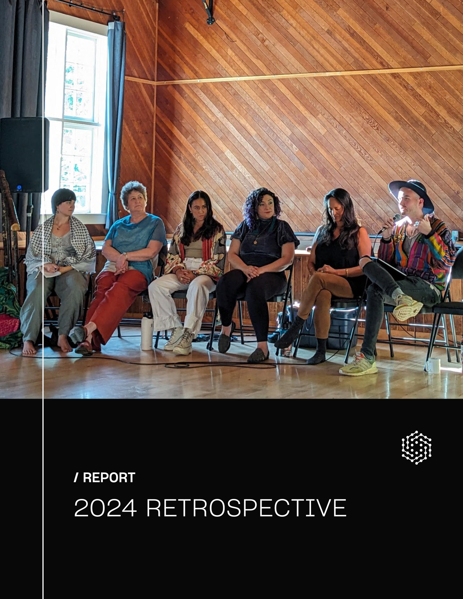 Panel of six women sitting in chairs during a discussion or presentation in a room with wooden walls, with one woman speaking into a microphone and wearing a colorful striped shirt and a large hat.