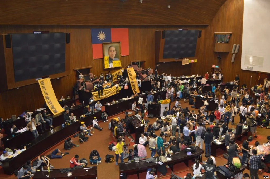 Large indoor gathering with many people seated and standing around tables, with prominent banners and a portrait of a man on the wall, indicating a political event in Taiwan.