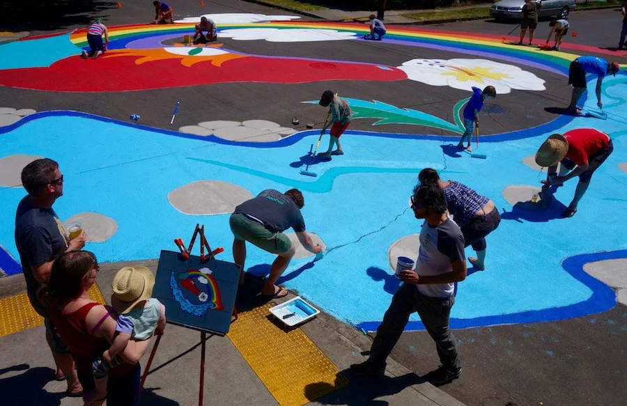 Group of people painting a colorful mural on a sidewalk with rainbows, clouds, and a blue sky.