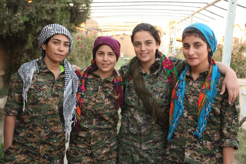 Four women dressed in camouflage military uniforms standing together outdoors with their arms around each other.
